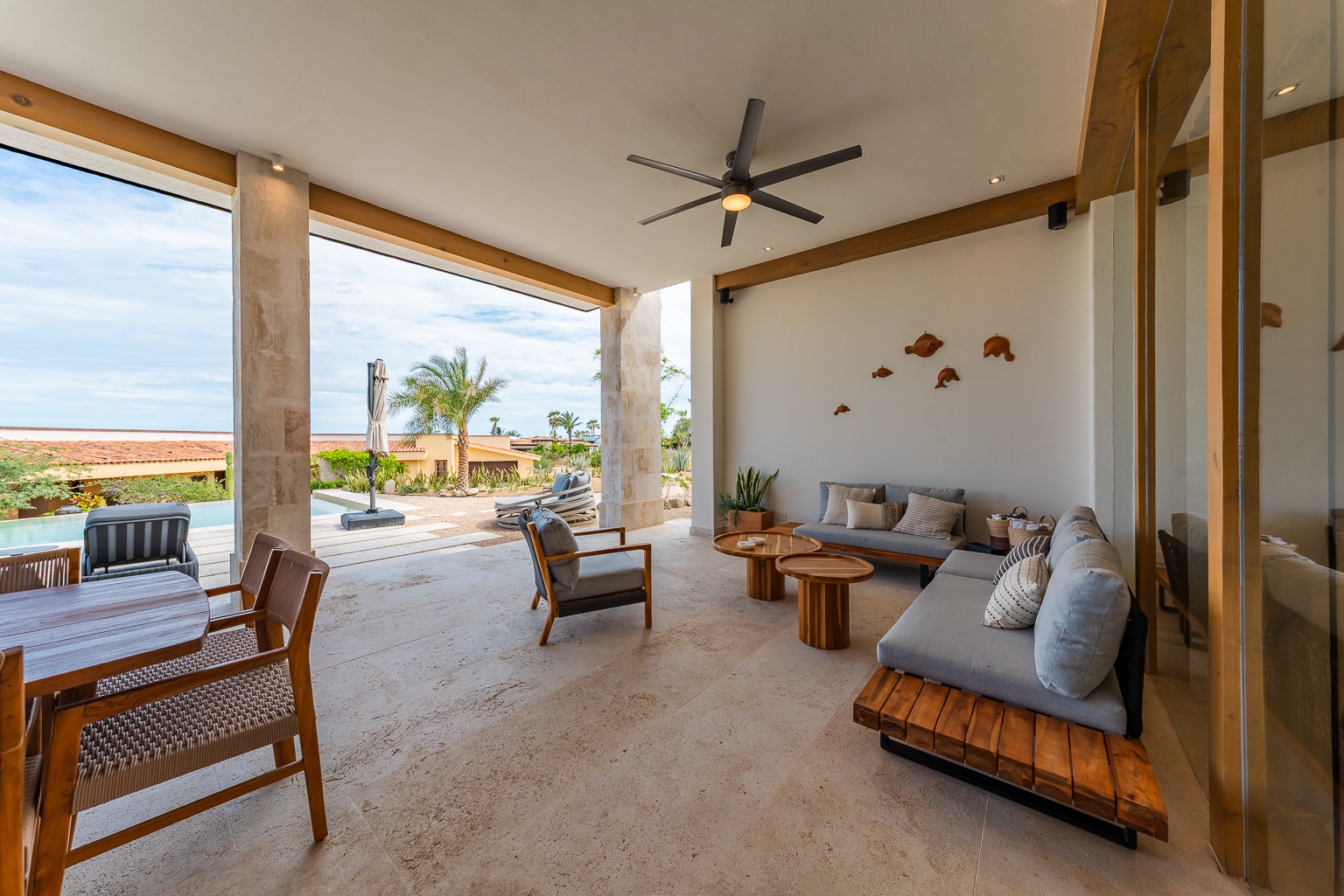 Stylish semi-open lounge with modern furnishings and a refreshing ceiling fan opens to pool views and lush tropical palms. Unwind here in natural coastal elegance.