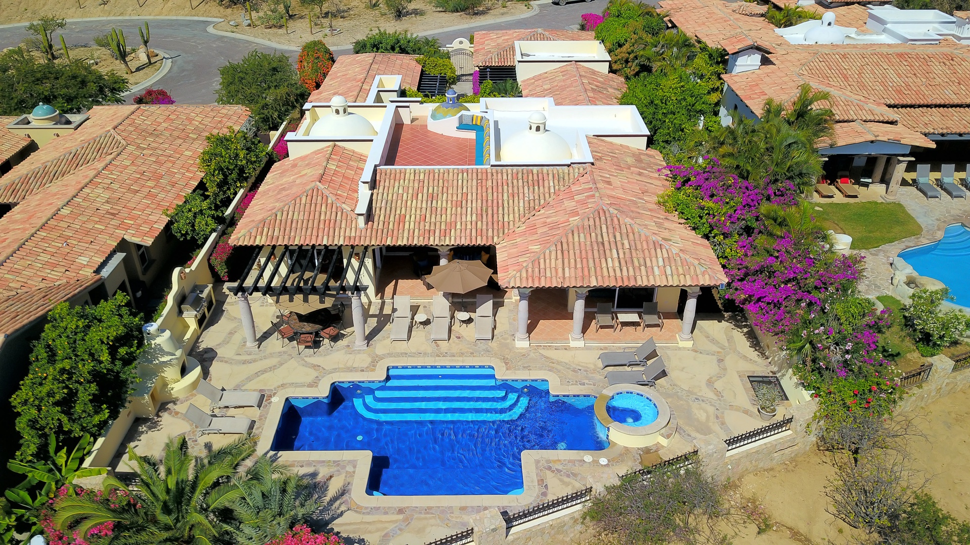 Stunning aerial view of a desert villa featuring a multi-level swimming pool with spa, clay tile roofing, and lush landscaping amid the natural surroundings.
