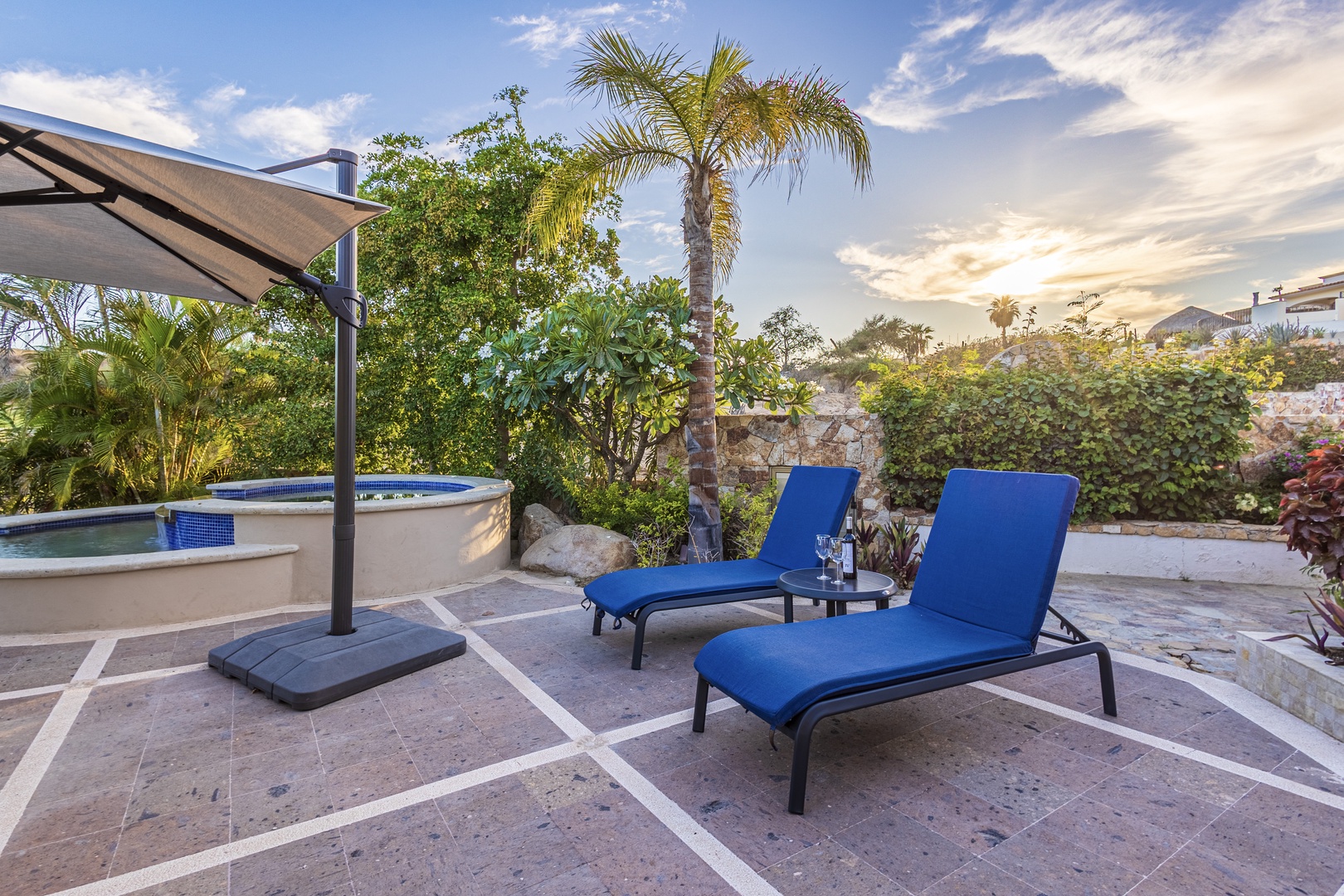 Unwind on your private patio with two lounge chairs beside a sparkling hot tub, surrounded by lush tropical palms and vibrant gardens under warm, golden skies.