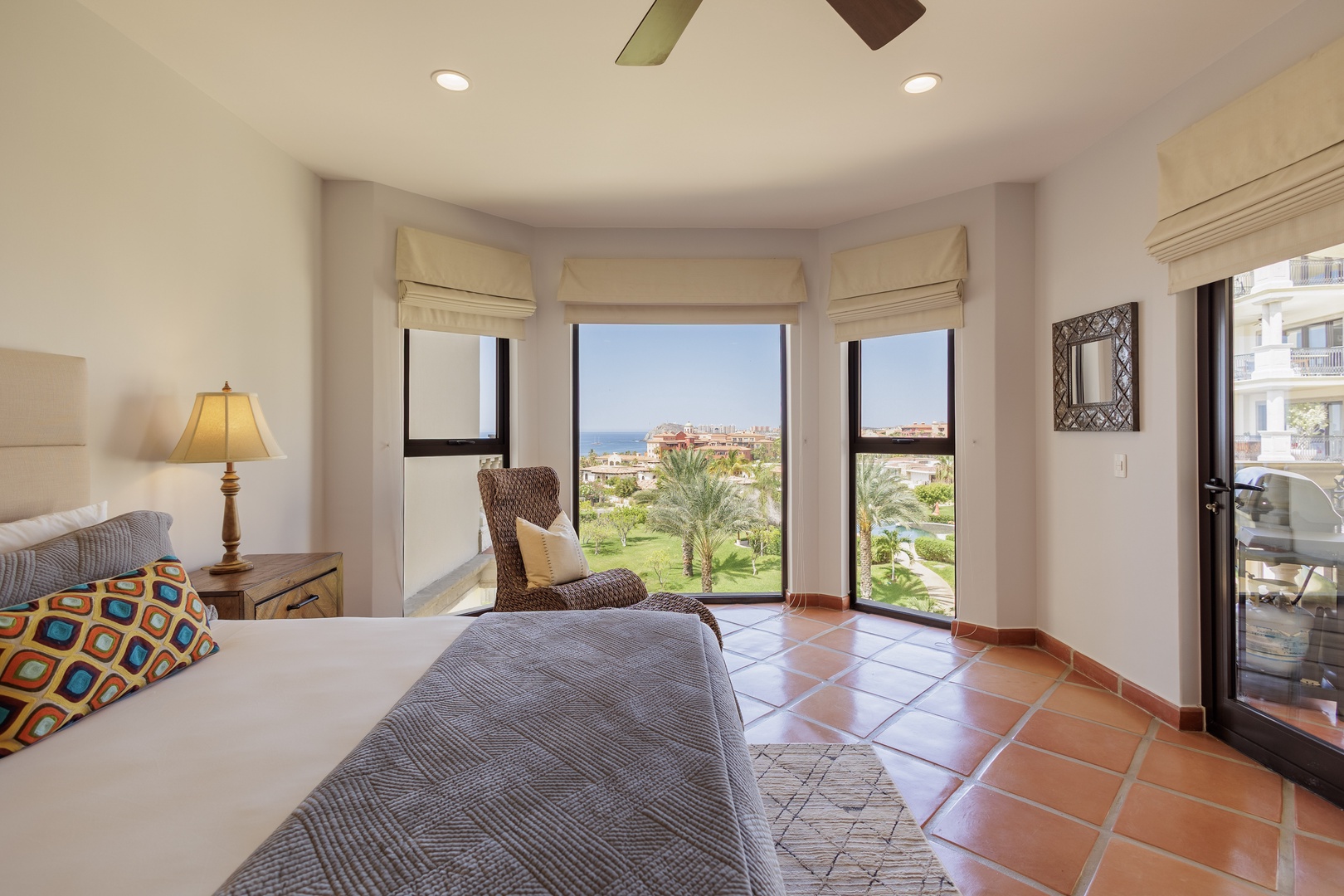 Unwind in your bright bedroom featuring a cozy wicker reading chair, ceiling fan, and breathtaking ocean and garden views through expansive windows.