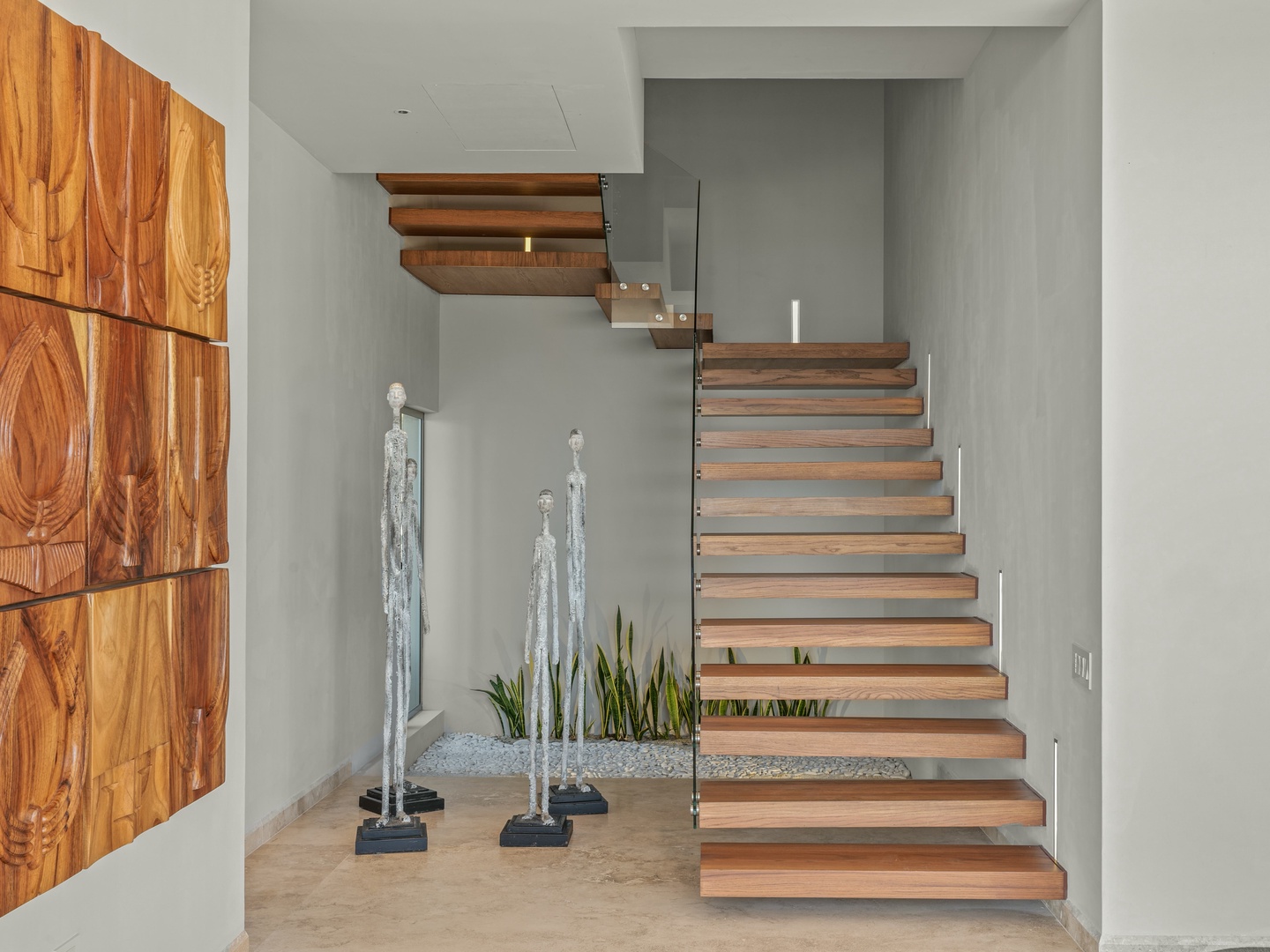 A striking modern staircase with floating wood treads and sleek glass railing welcomes you into this stylish space. Note the artistic sculptures and vibrant greenery adding character to your entry.