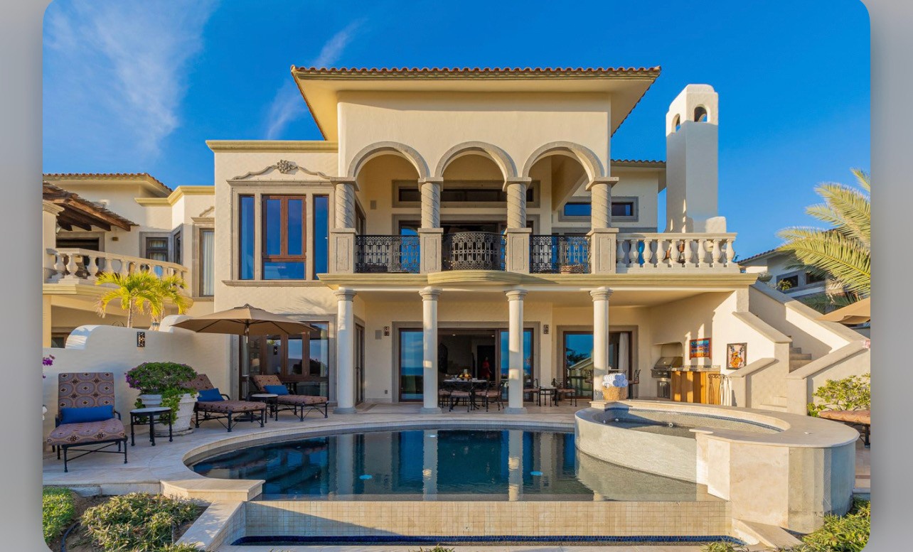 Elegant Mediterranean villa with grand arched balconies, ornate railings, and classical columns. Stunning outdoor space features a pristine pool surrounded by stylish seating, lush landscaping, and endless blue skies.