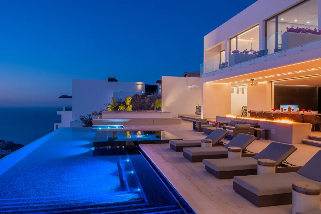 A contemporary property featuring multiple levels, illuminated pool areas, and outdoor lounging spaces overlooking the surrounding landscape at twilight.