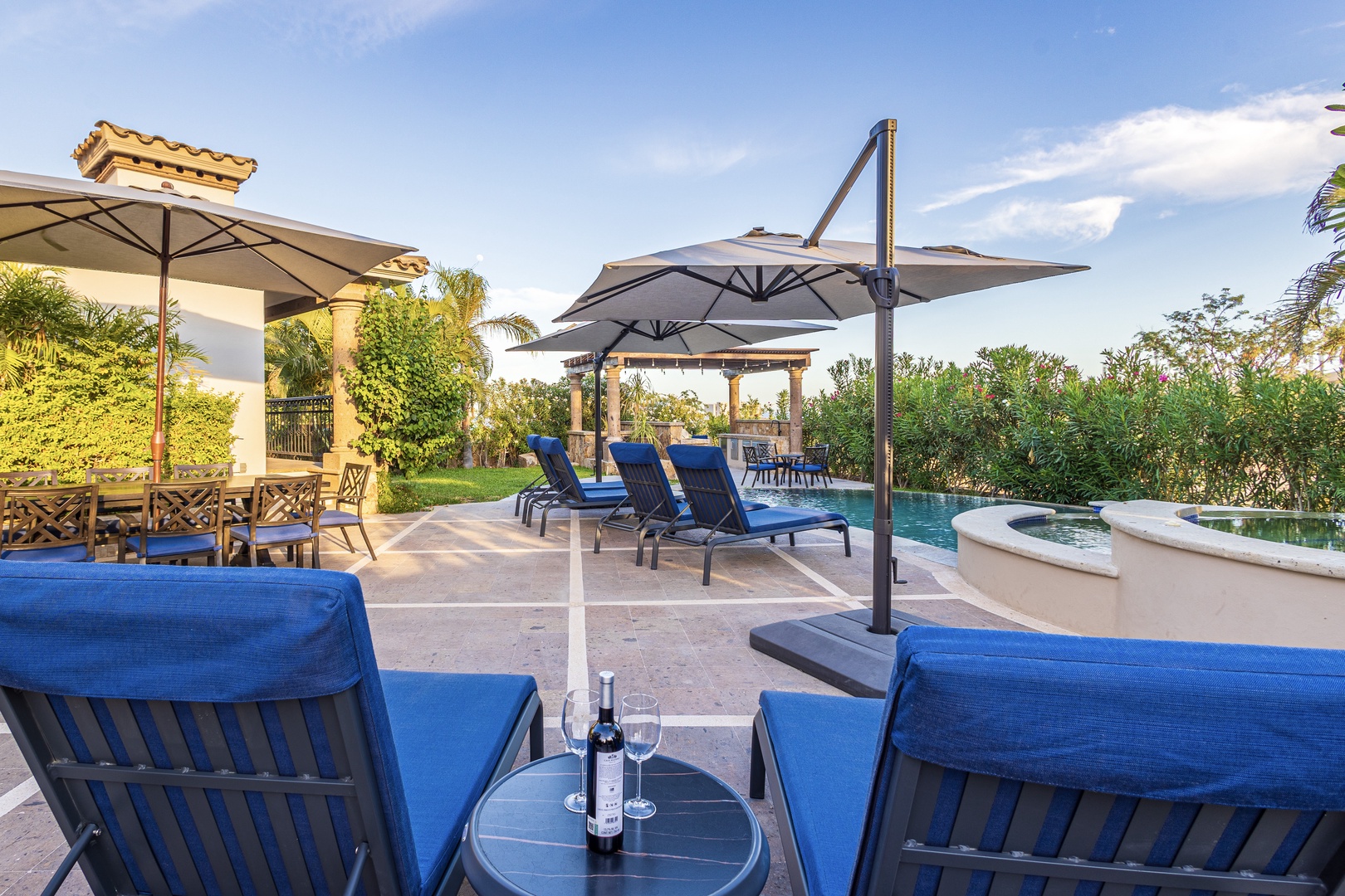 A sun-drenched patio with plush blue loungers, shaded dining areas, and a sparkling pool set in a lush garden—your perfect spot to unwind with a glass of wine.