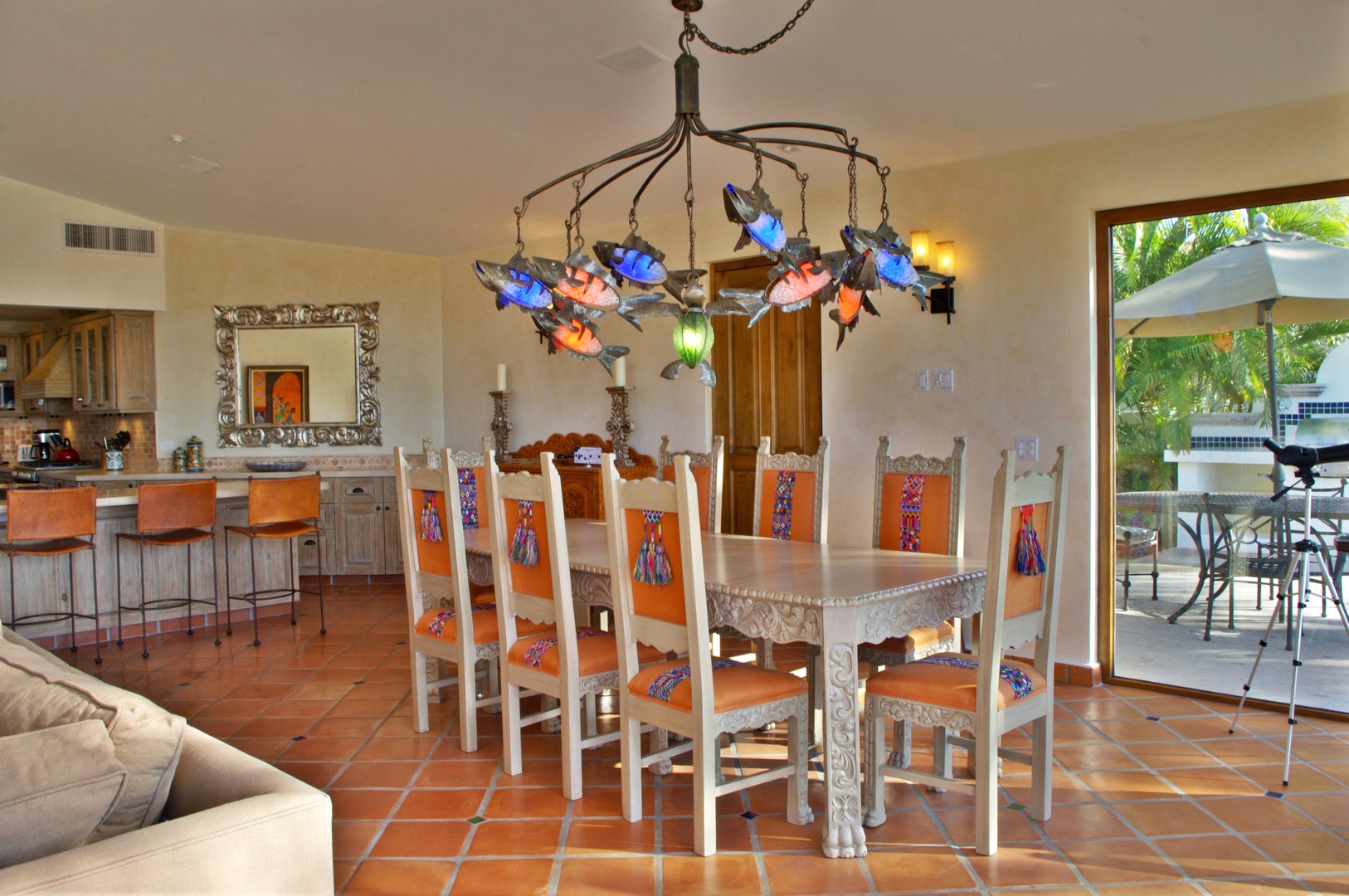 This spacious, sun-drenched dining area invites you to gather around a stunning handcrafted table beneath a vibrant hand-blown glass chandelier, with tropical views spilling in through open patio doors.