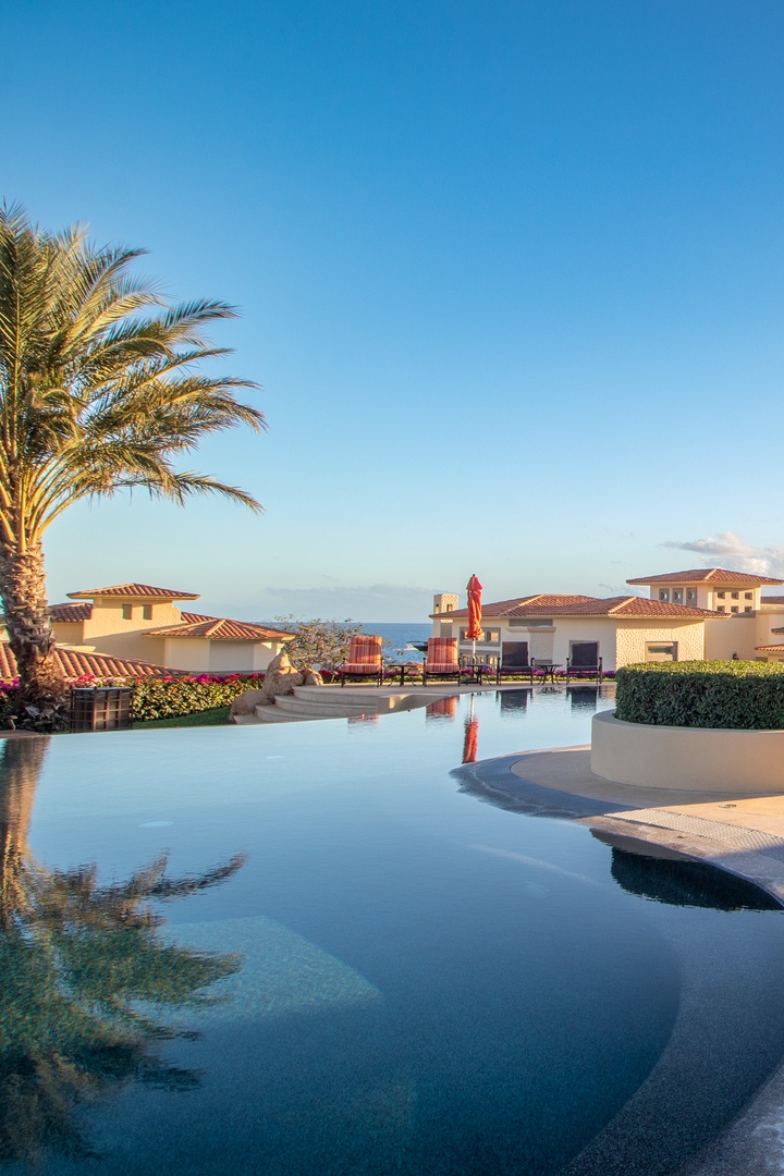 Serene infinity pool curves through lush landscaping, with terracotta-roofed villas nestled against the coastline under a golden sunset sky.