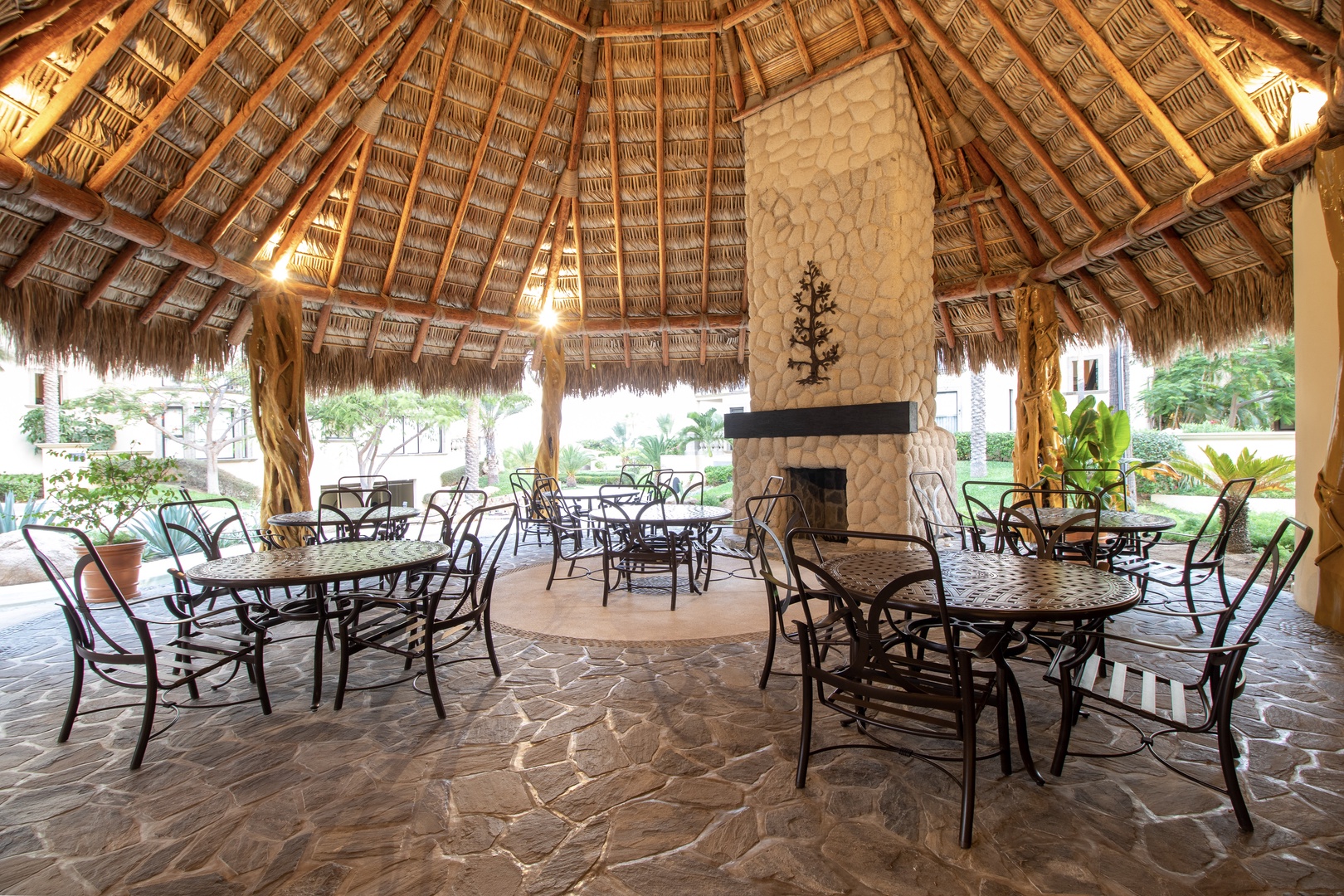 A stunning palapa-covered gathering space with a stone fireplace and elegant dining tables—perfect for shared meals, lively conversations, or simply savoring the tropical garden views surrounding you.