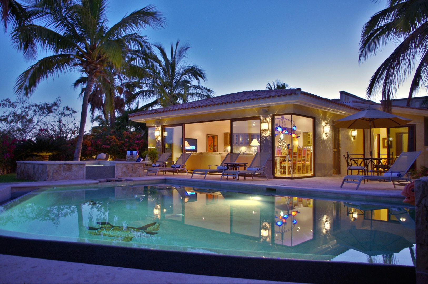 A stunning tropical villa with glowing warm lights and shimmering pool reflects the twilight sky, surrounded by swaying palms and lush gardens in this serene Caribbean-inspired setting.