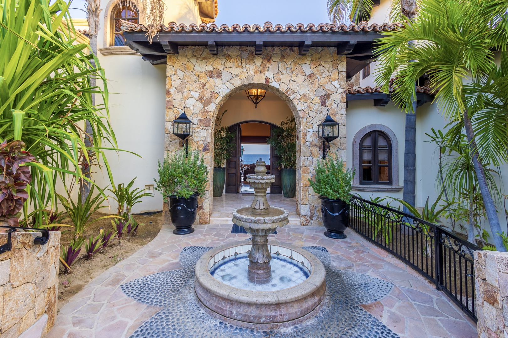 A grand stone archway frames your entrance, where a tiered fountain welcomes you with the soothing sound of flowing water, surrounded by lush tropical palms and Mediterranean-inspired architecture.