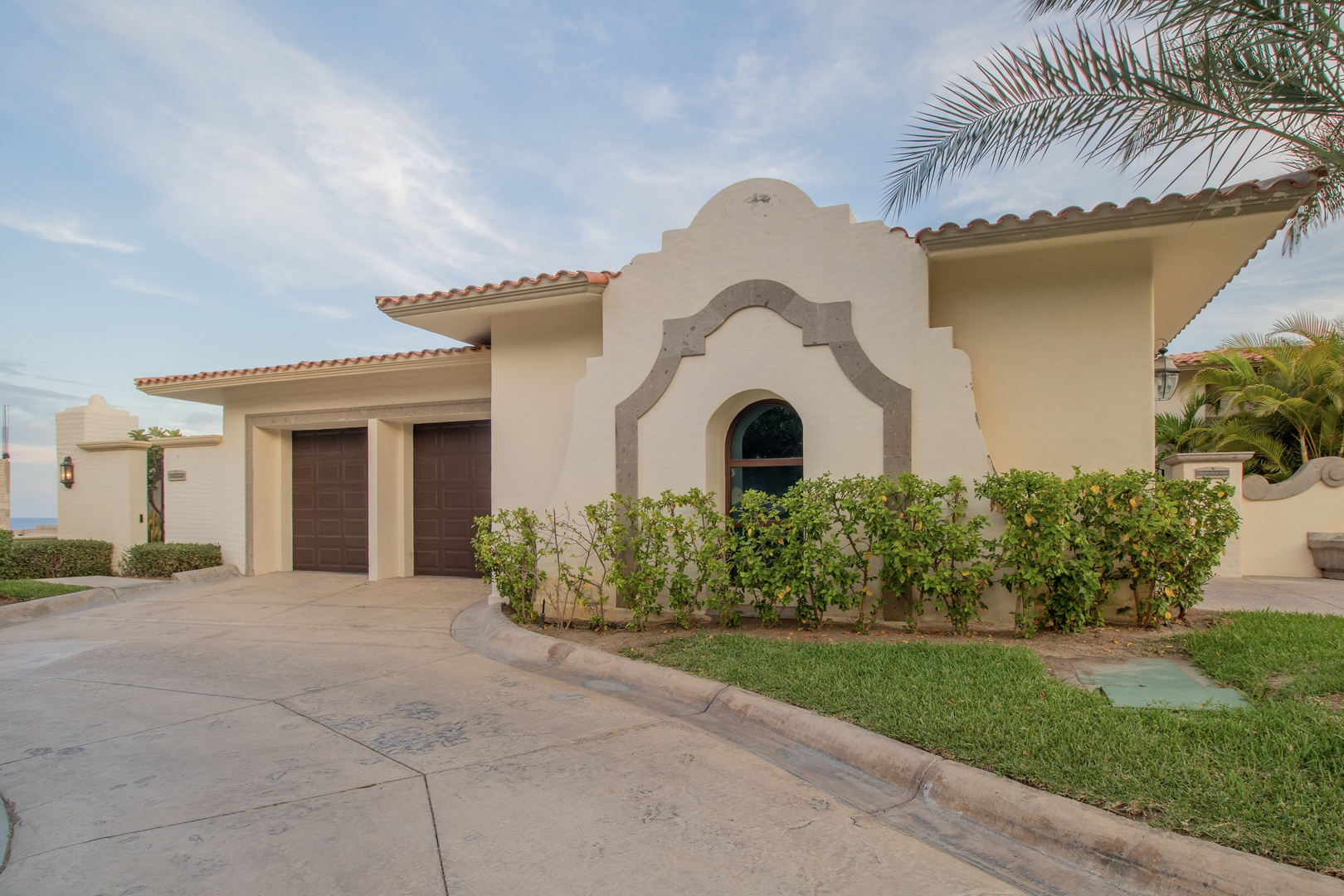 Charming Spanish-style villa entrance welcomes guests with Mediterranean architectural details, two-car garage, manicured landscaping, and elegant terra cotta tile roofing set against a tranquil coastal backdrop.