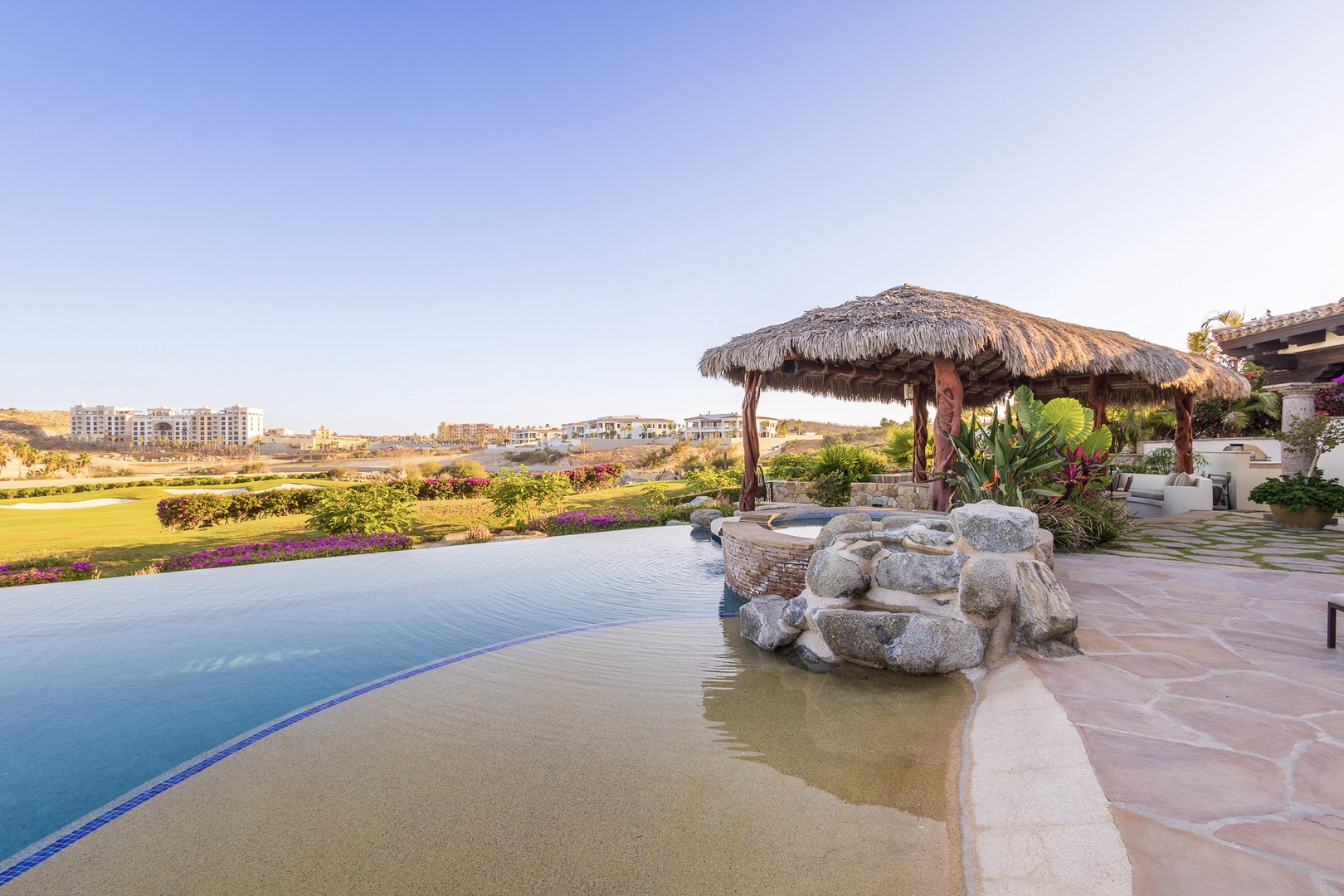 A stunning outdoor oasis featuring an infinity pool overlooking lush golf greens, complete with a thatched palapa and natural stone hot tub surrounded by tropical gardens and flowering plants.