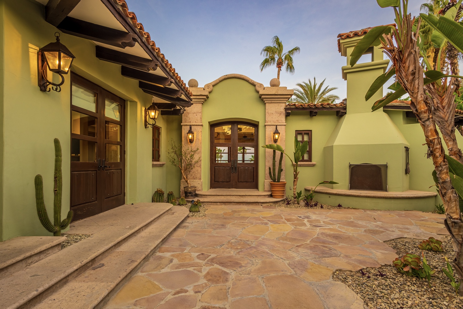 A charming Mediterranean-style entrance welcomes guests with warm tile roofing, arched doorways, and lush tropical landscaping framed by swaying palms and desert plants.