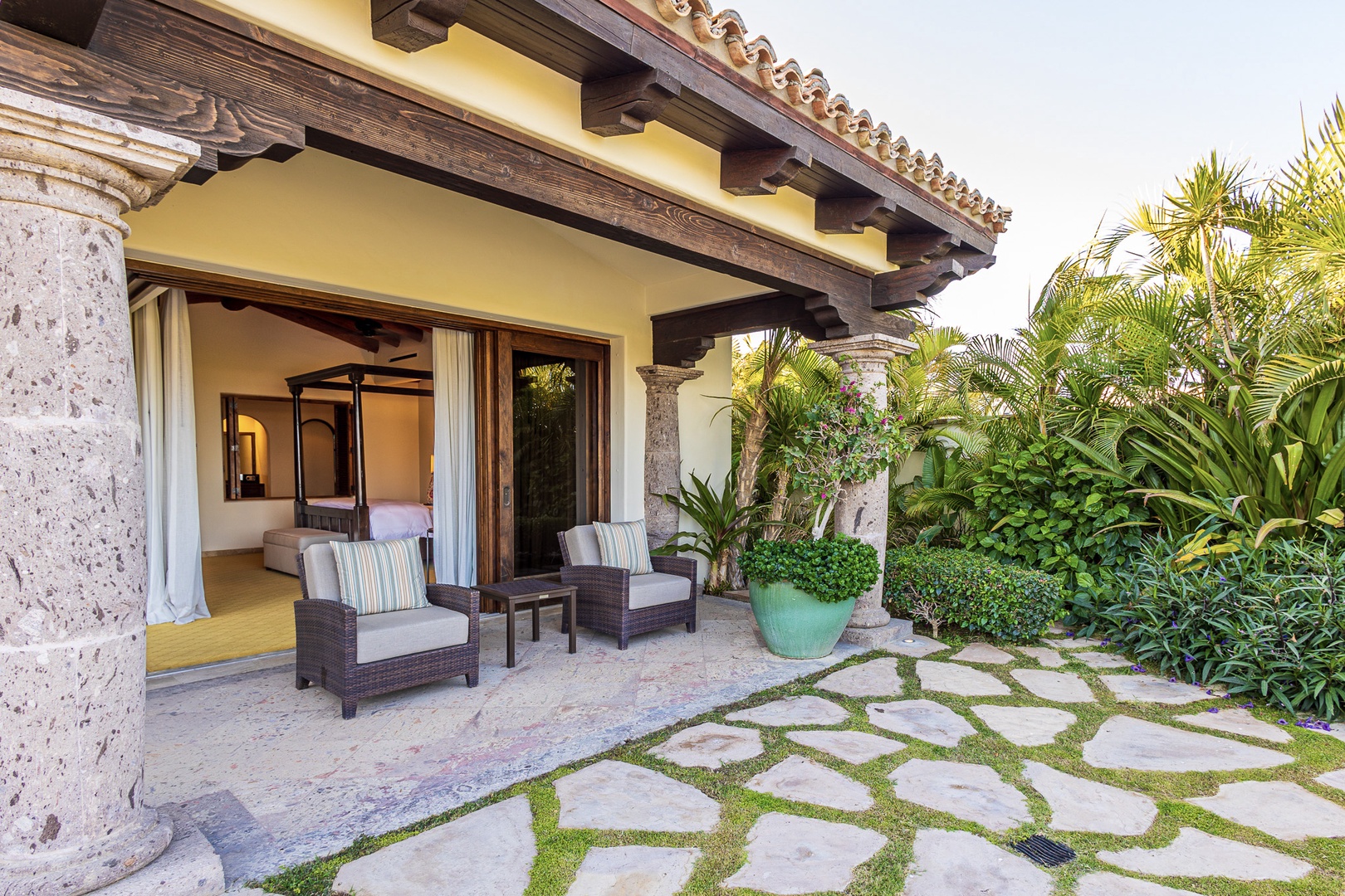 A stone-paved path winds through lush tropical gardens, leading to a private terrace featuring an elegant architectural design with exposed beam ceilings and comfortable seating overlooking the surrounding natural landscape.