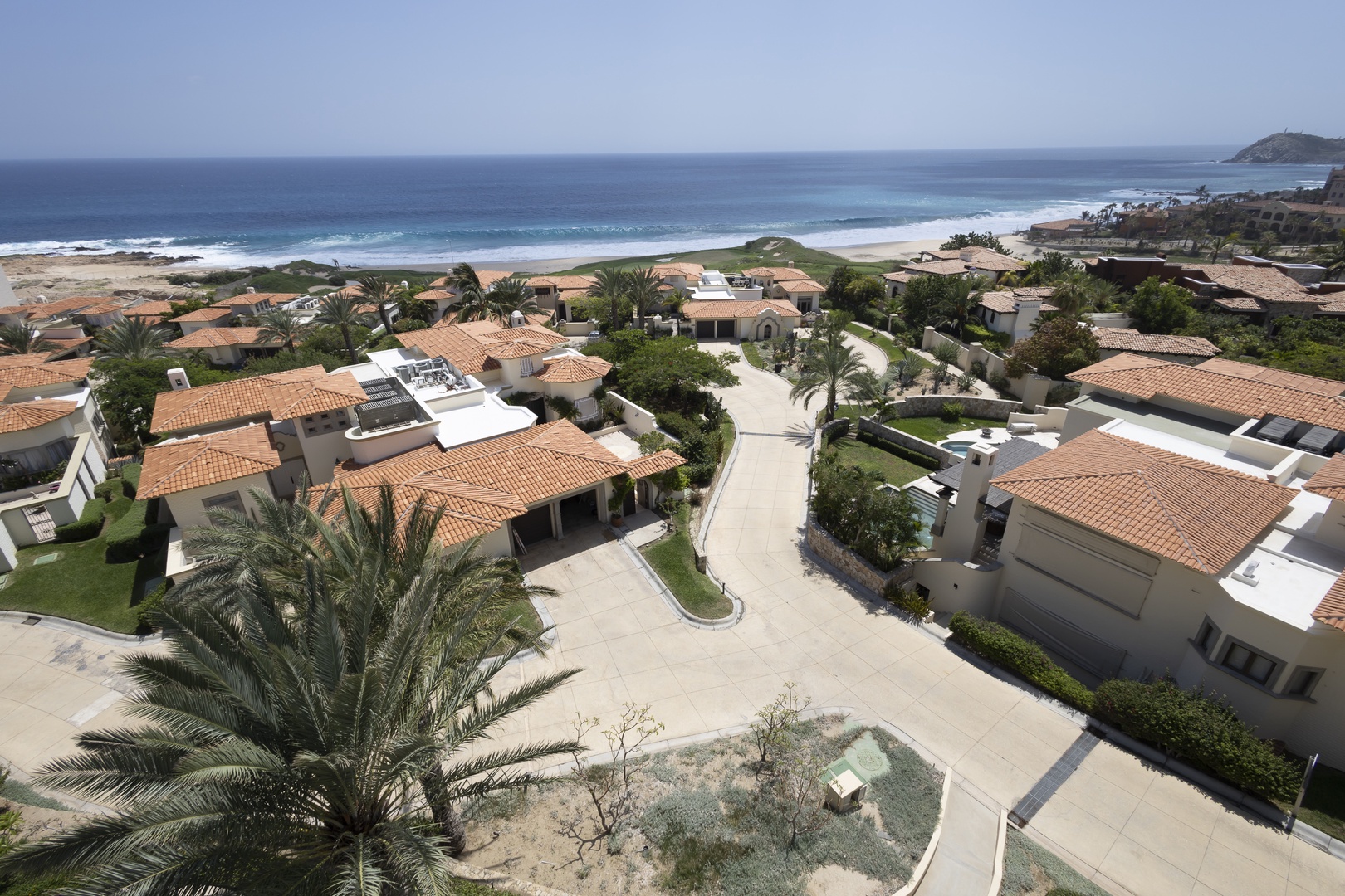 Elevated view of a beachfront resort community with terracotta-roofed villas, palm-lined streets, and pristine coastline stretching along the horizon.