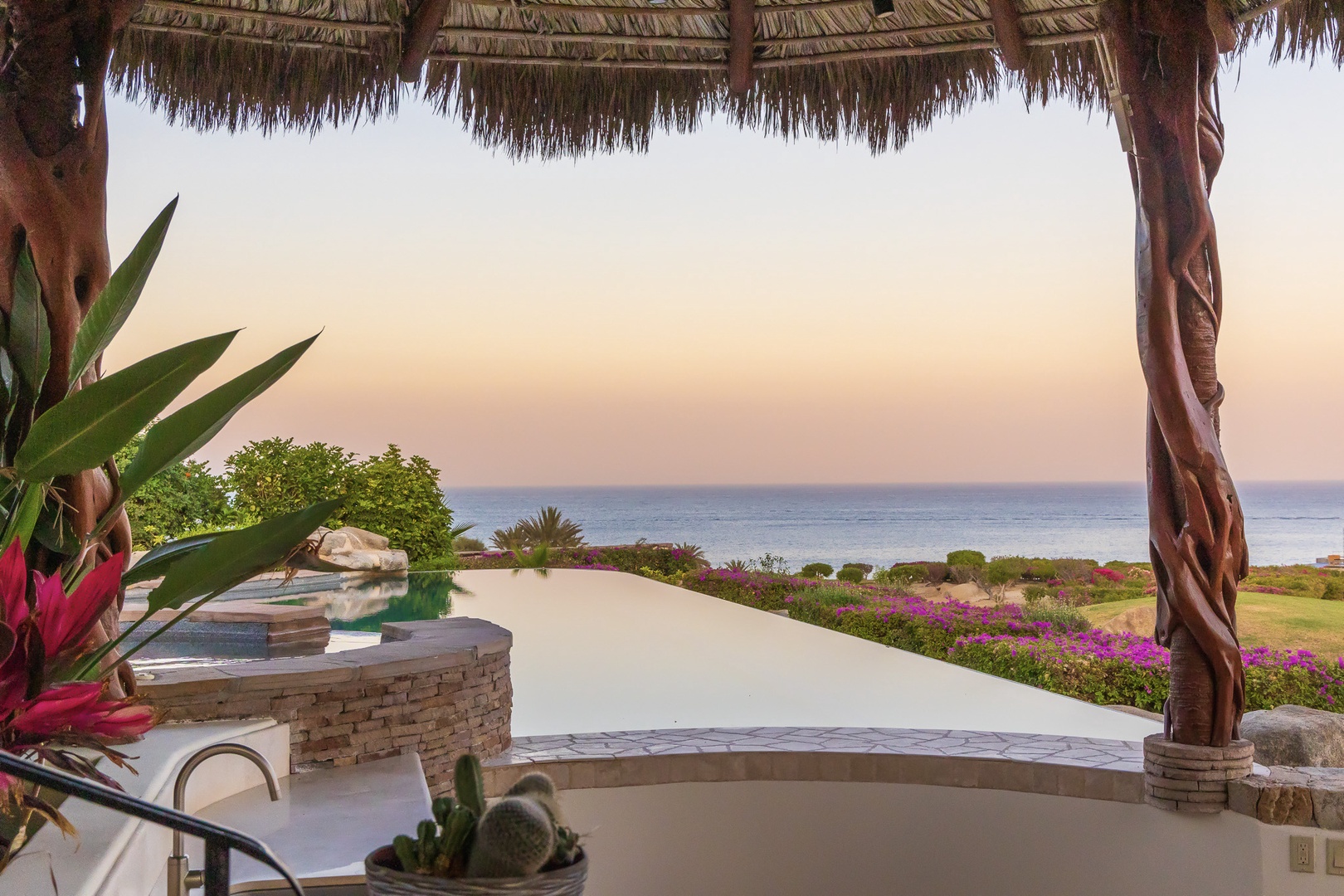 Private infinity pool overlooks the sea at sunset, surrounded by vibrant tropical gardens and sheltered beneath a traditional palapa—a stunning backdrop to the coastal landscape.