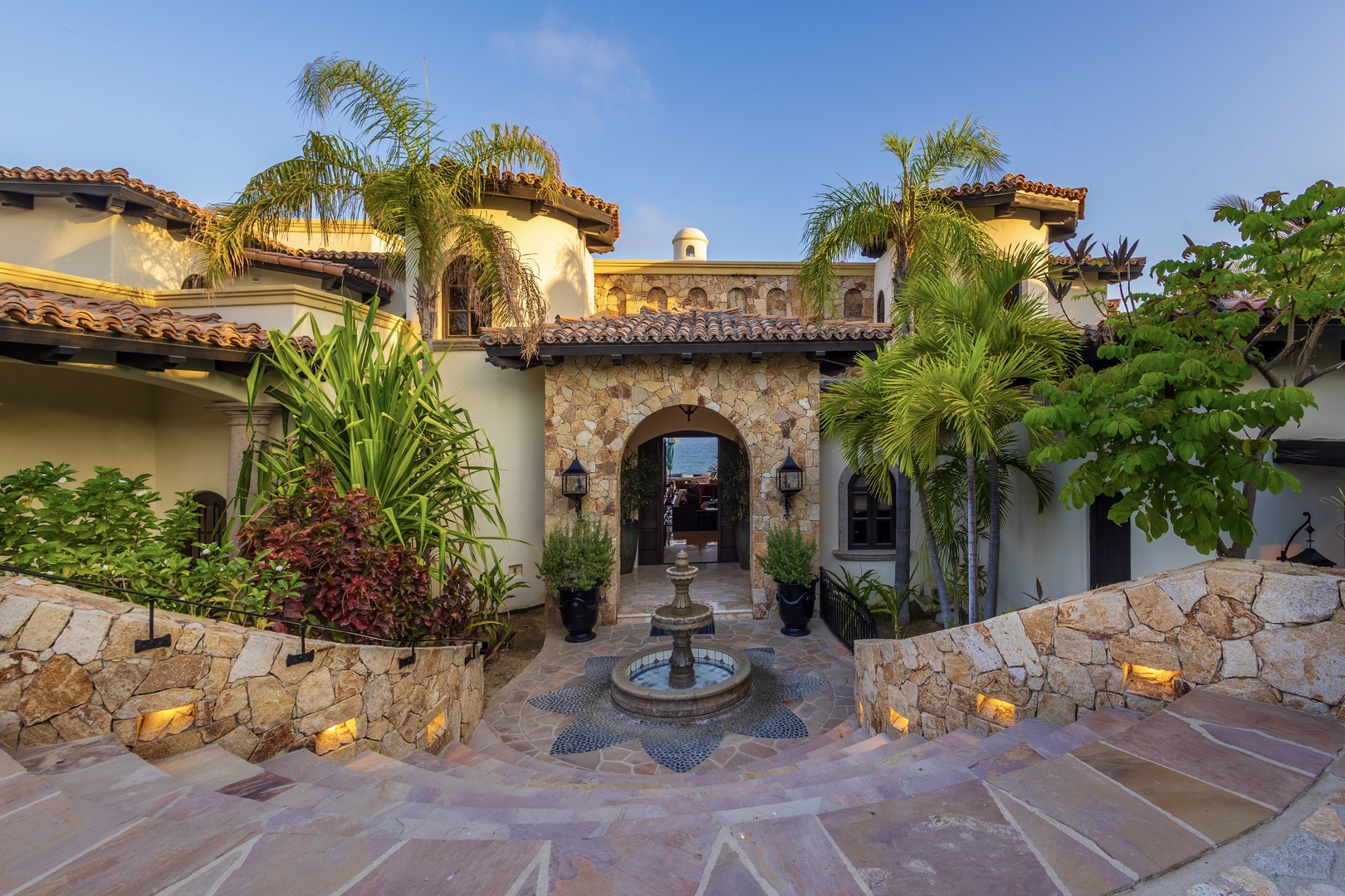 A stunning Mediterranean-style villa entrance features an arched stone gateway, courtyard fountain, and lush tropical landscaping with ambient lighting at dusk.