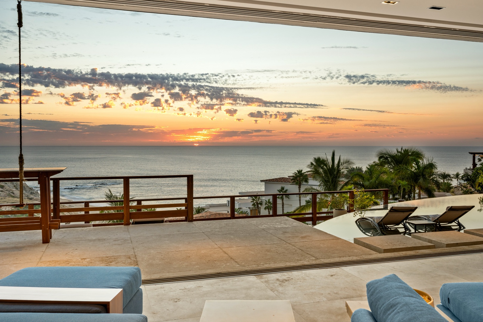 Stunning oceanfront terrace overlooking palm-lined coastline at sunset, framed by the covered lounge area with modern seating and recliners for relaxing at day's end.
