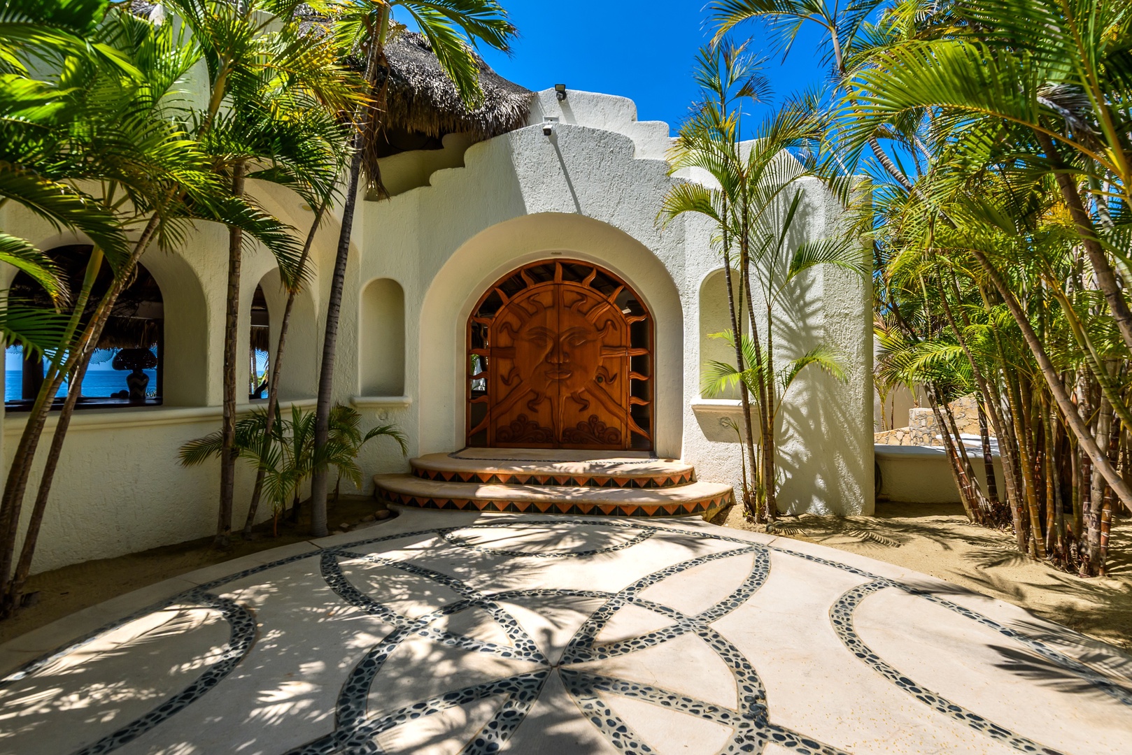 A stunning beachside villa entrance framed by tropical palms and swaying fronds, featuring distinctive architecture with carved wooden doors, tiled steps, and an artful pebbled courtyard welcoming you to paradise.