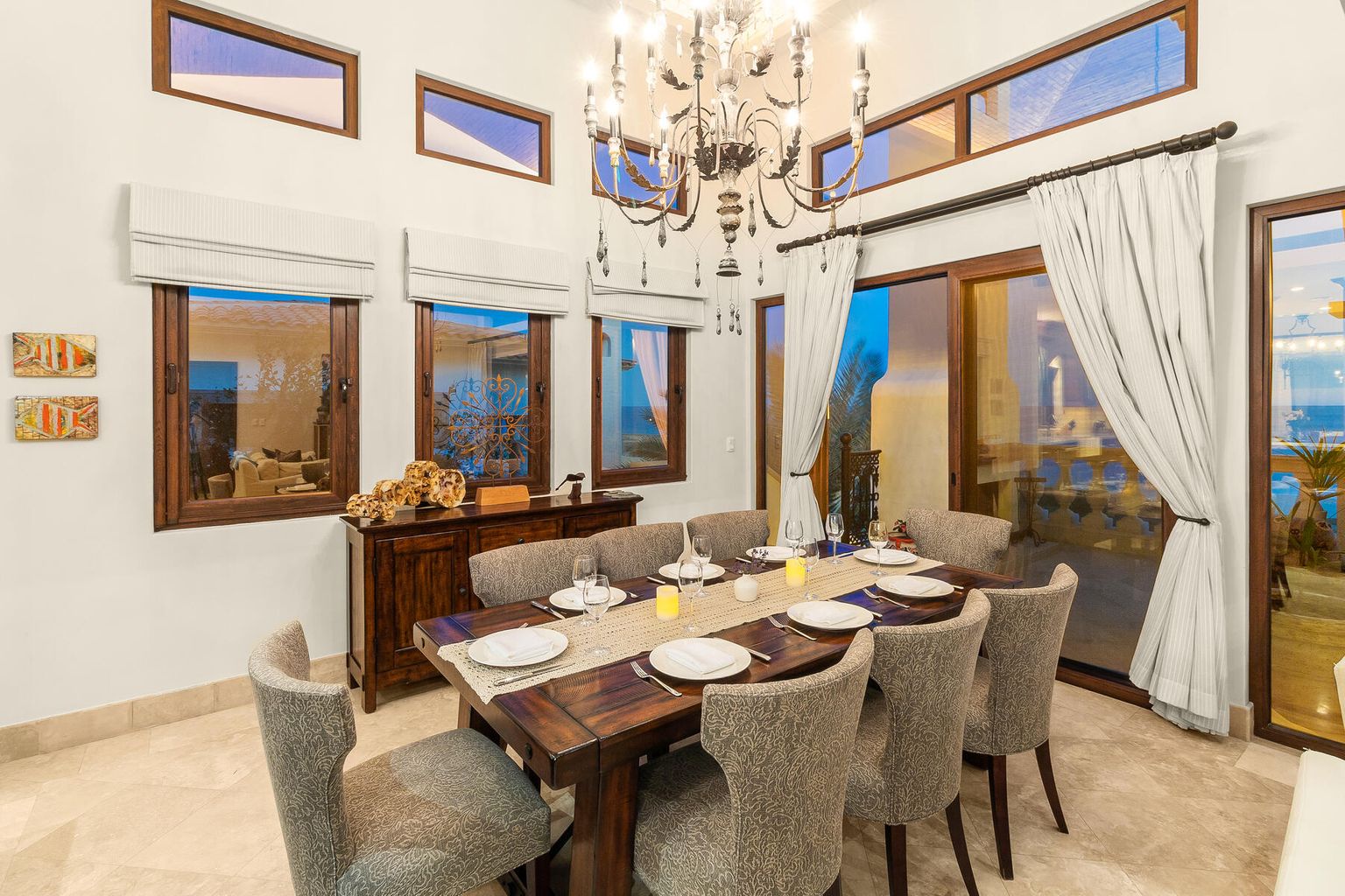 Gather at your elegant dining table beneath soaring ceilings and a statement chandelier, with windows framing serene ocean views beyond.