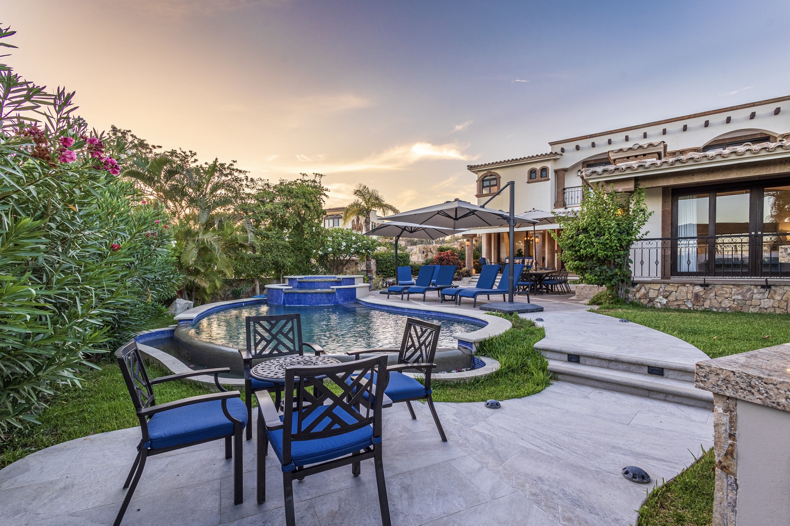 Luxury vacation villa with private pool, lush tropical gardens, and multiple outdoor lounging areas framed by elegant Spanish-style architecture at sunset.