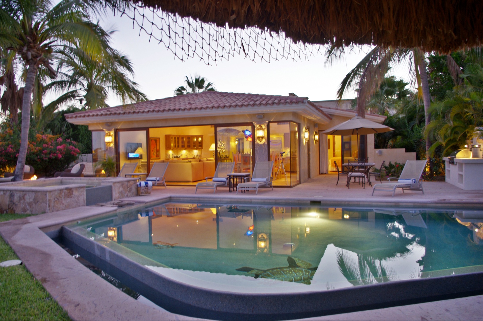 A tropical villa with terracotta roof sits amid lush palms, featuring a thatched palapa over the sparkling pool and illuminated outdoor living spaces at twilight.
