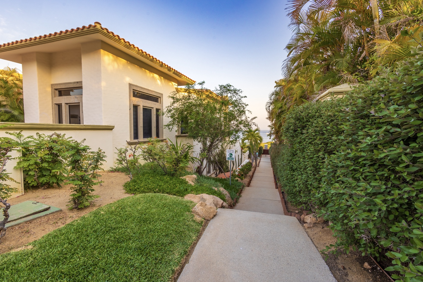 A welcoming pathway lined with lush greenery and palm trees leads to the property's charming entrance, framed by natural landscaping and a terra-cotta-tiled roof.