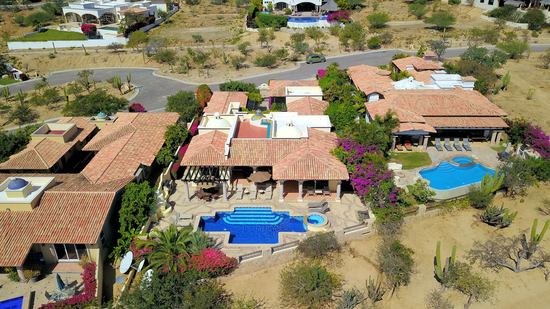 Aerial view of a sprawling villa complex with distinctive red tile roofs, multiple swimming pools, and desert landscaping in a quiet residential neighborhood.