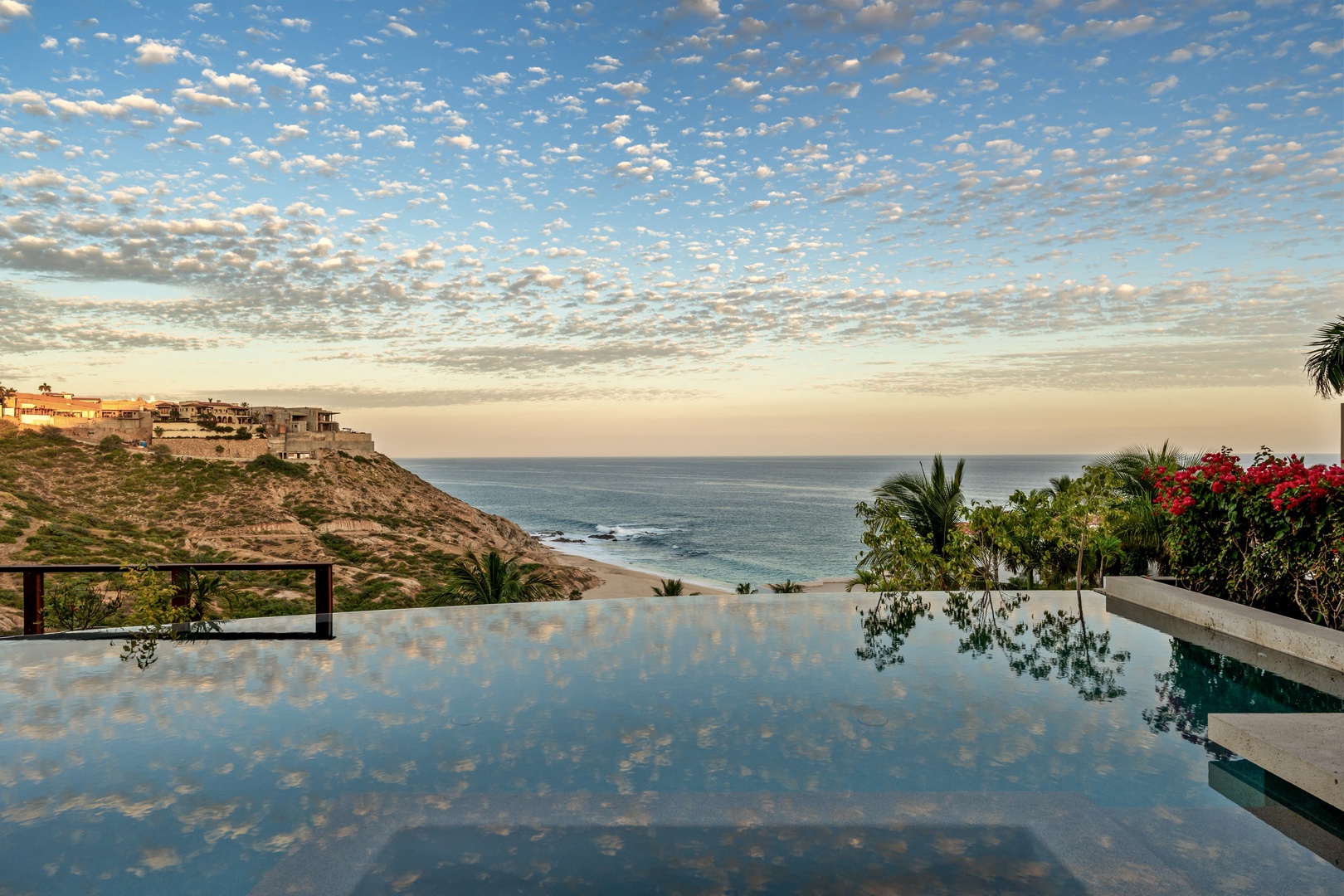 An infinity pool overlooks serene ocean waves and coastal cliffs, framed by lush tropical greenery and a dramatic sky painted with delicate clouds at golden hour.