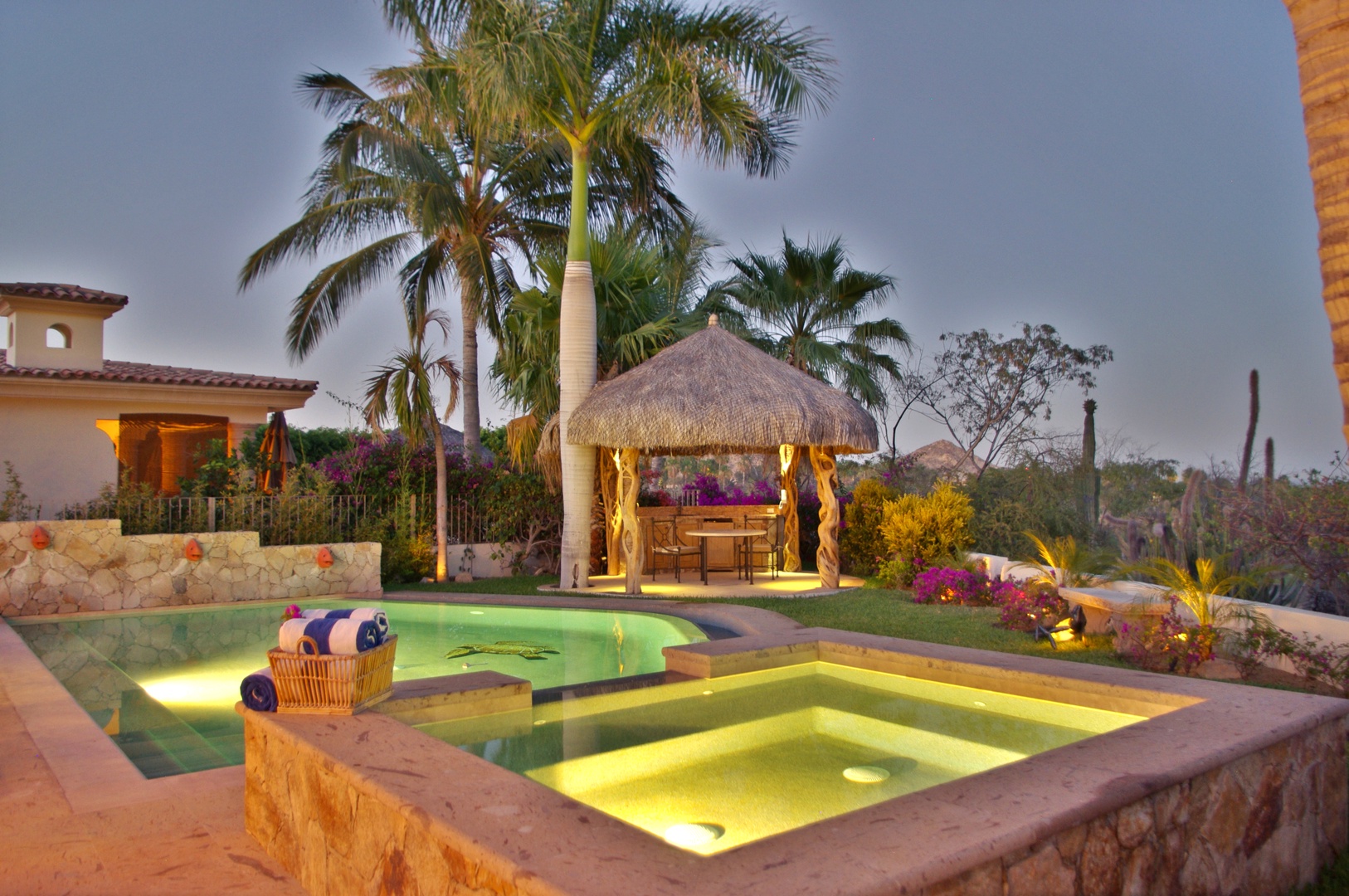 The illuminated pool and spa with thatched palapa create a private tropical oasis as dusk falls over this luxurious retreat.