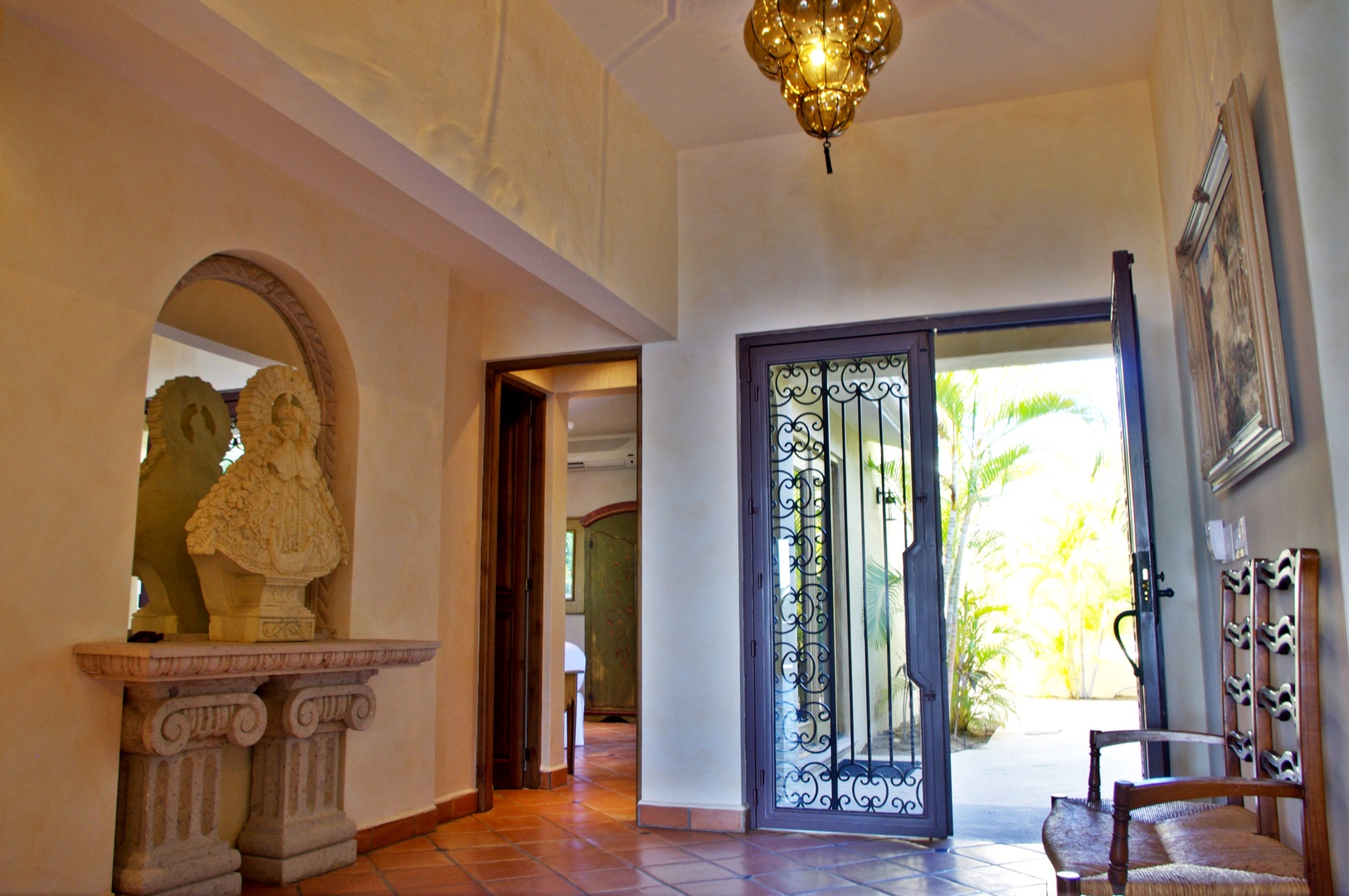 Step into an elegant entryway where intricate ironwork doors welcome natural light, revealing lush tropical greenery just beyond. Classic architectural touches and warm terracotta tiles set a refined, inviting tone.