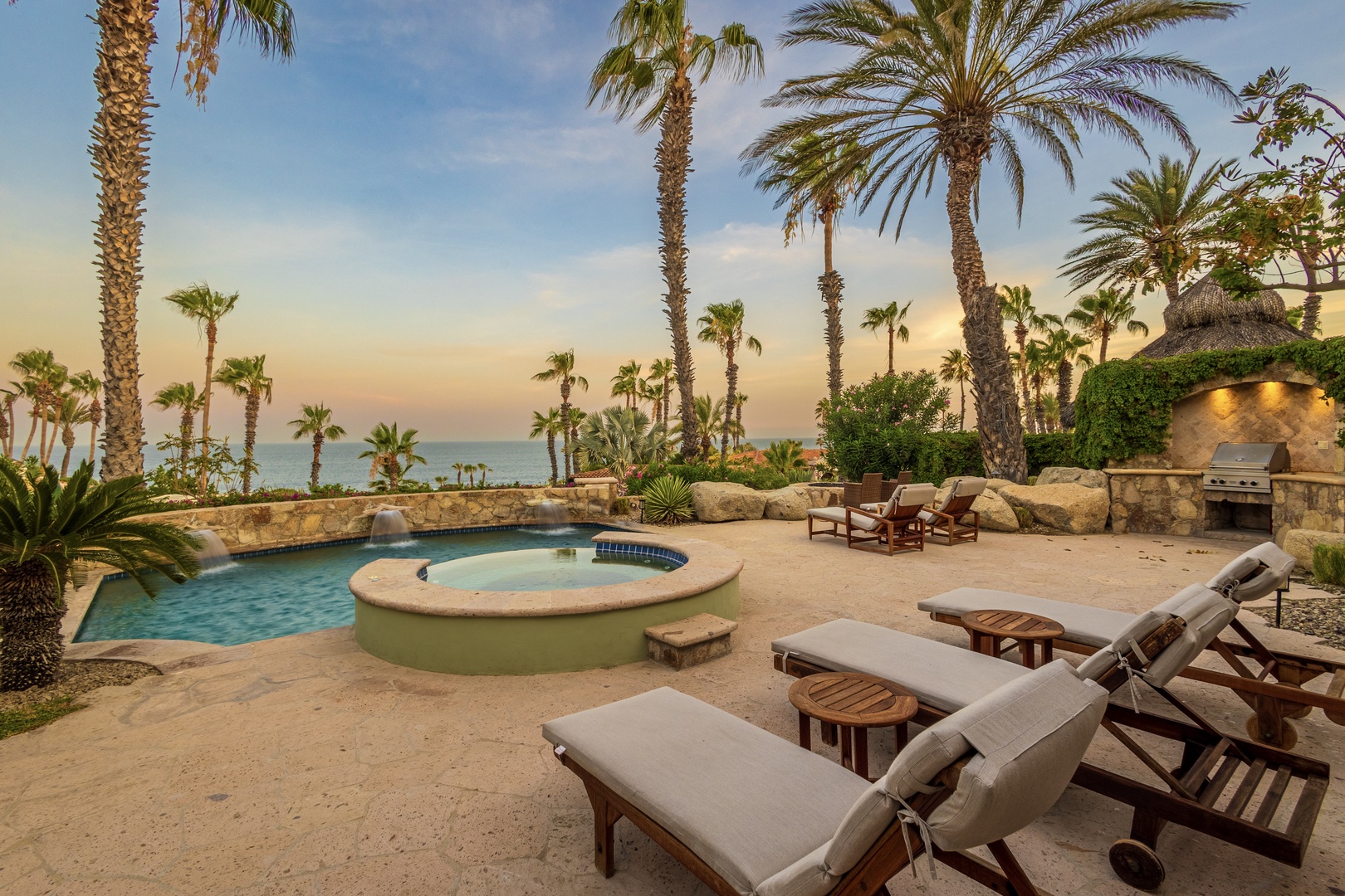 Beachfront oasis featuring swimming pool, hot tub, BBQ area, and ocean views framed by swaying palms at sunset. Lounge on the patio and soak in paradise.