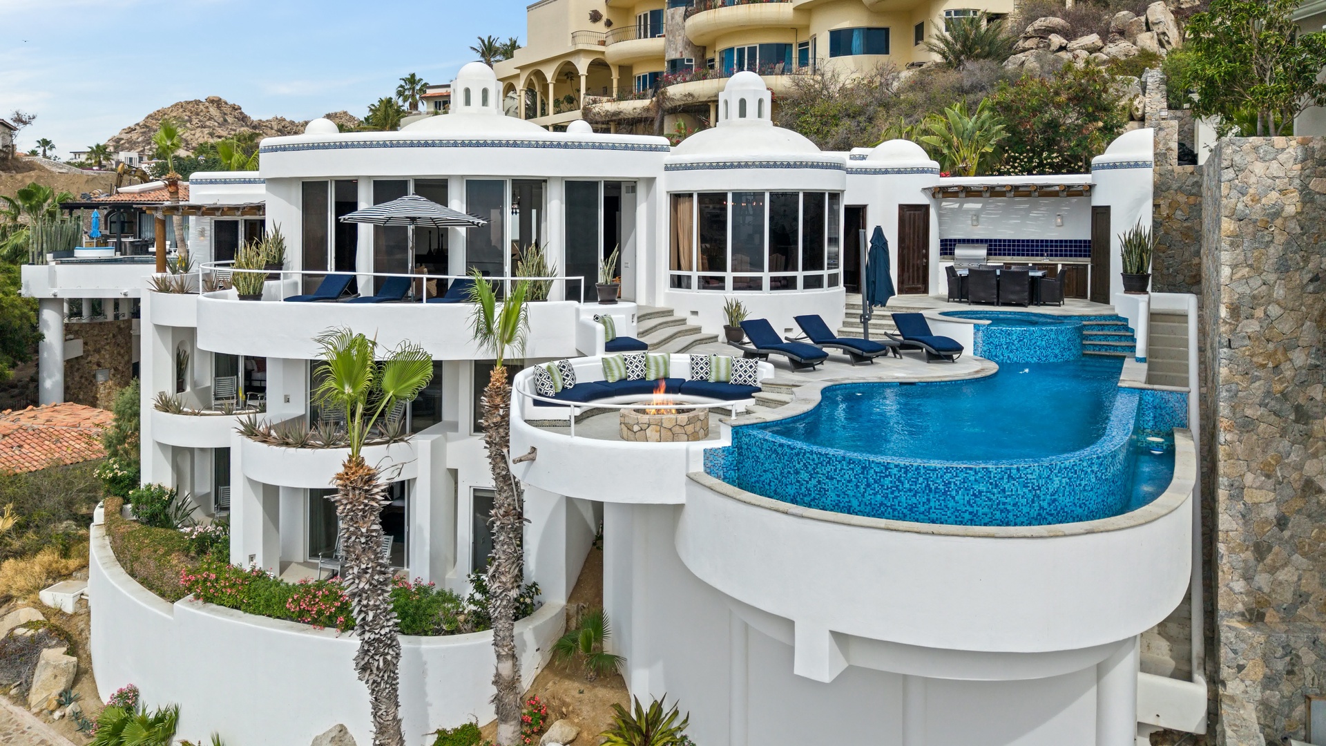 Stunning modern villa showcases pristine white architecture with a dramatic infinity pool cascading down terraced levels, surrounded by desert landscape and neighboring hillside homes.