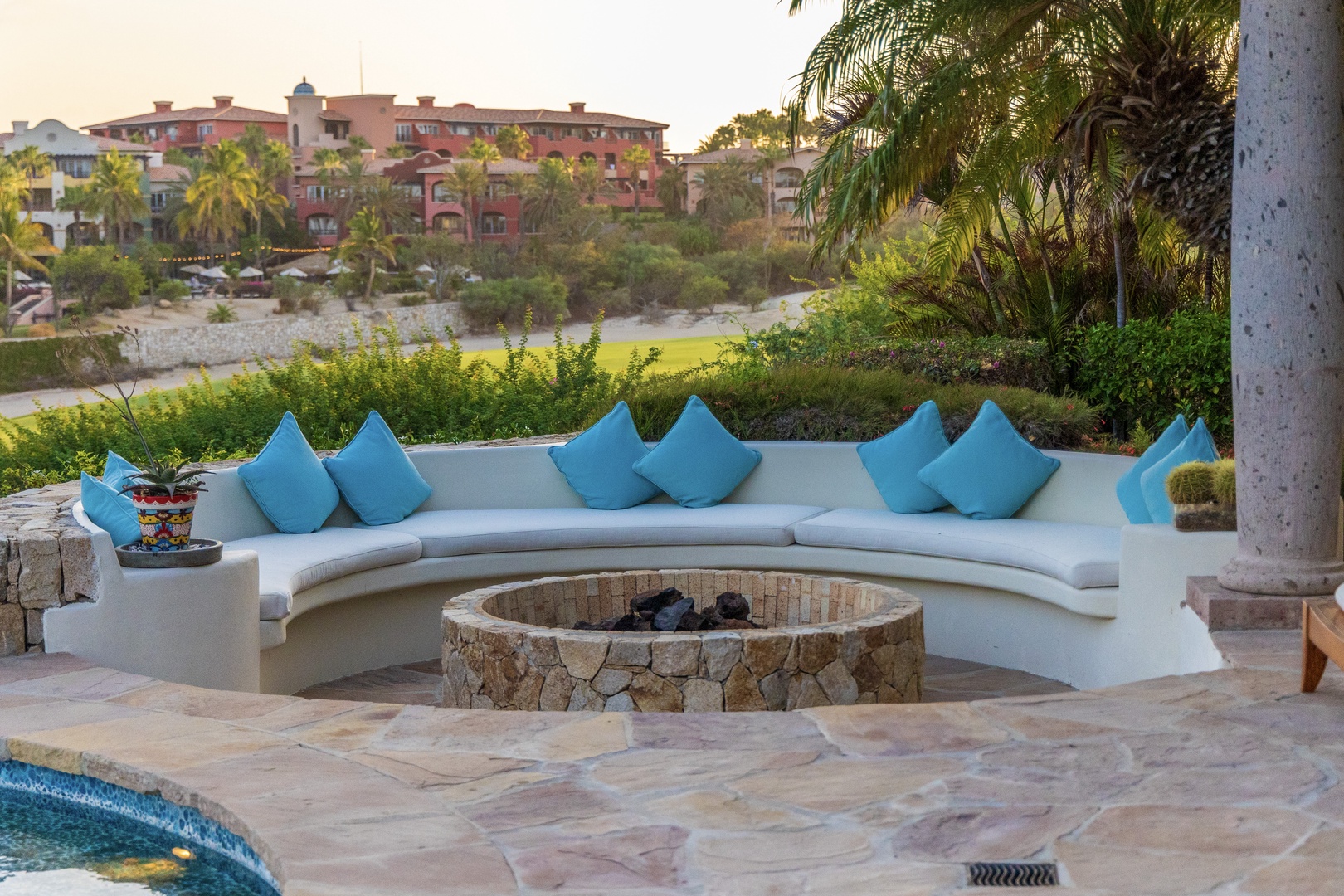 Settle into this inviting curved outdoor seating area with soft cushions and a central fire pit, perfect for sharing stories as the sun sets over the tropical landscape.