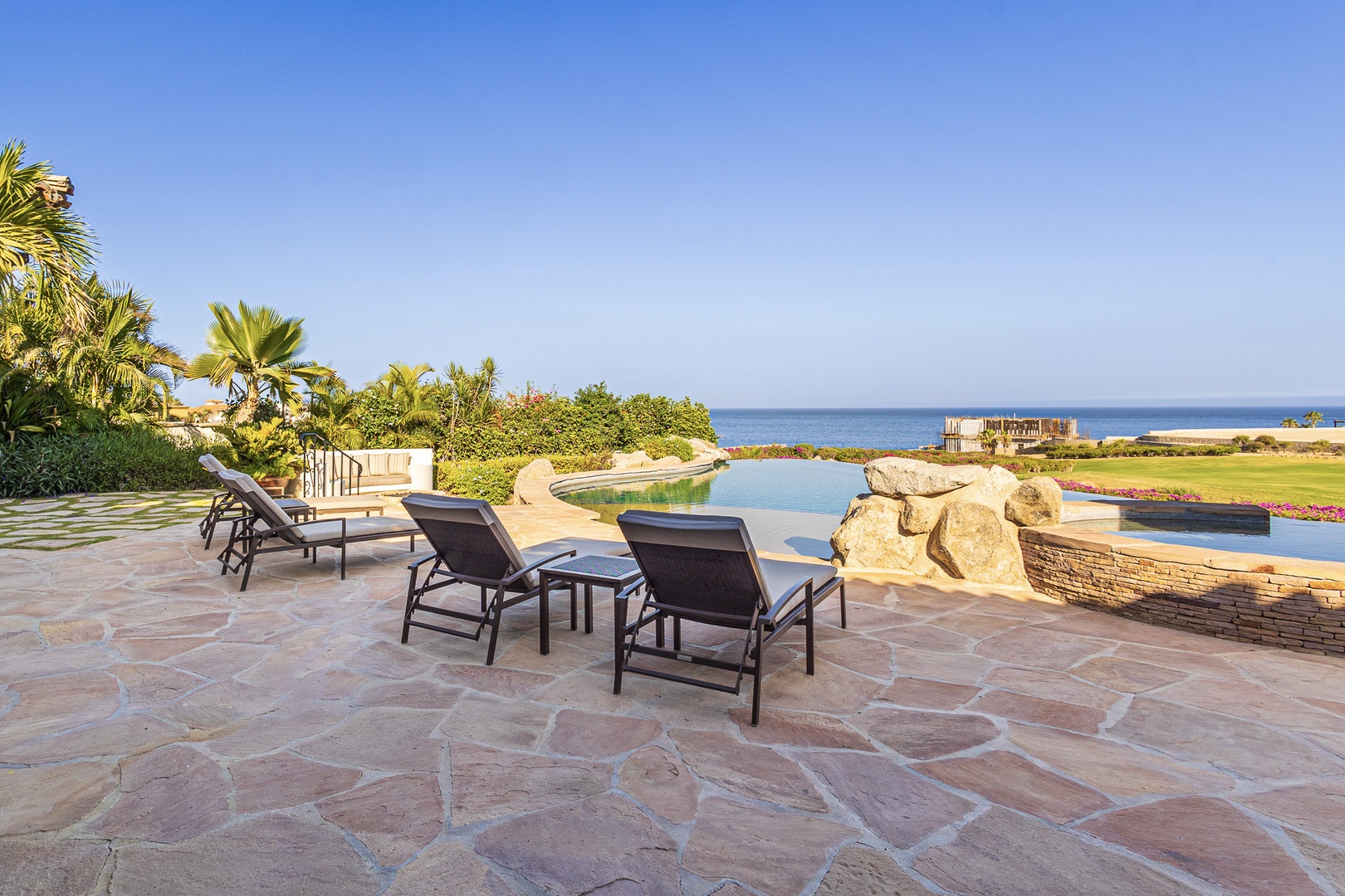 Expansive outdoor patio with infinity-edge pool overlooking tranquil coastline, lush tropical landscaping, and ocean horizon in Cabo San Lucas.