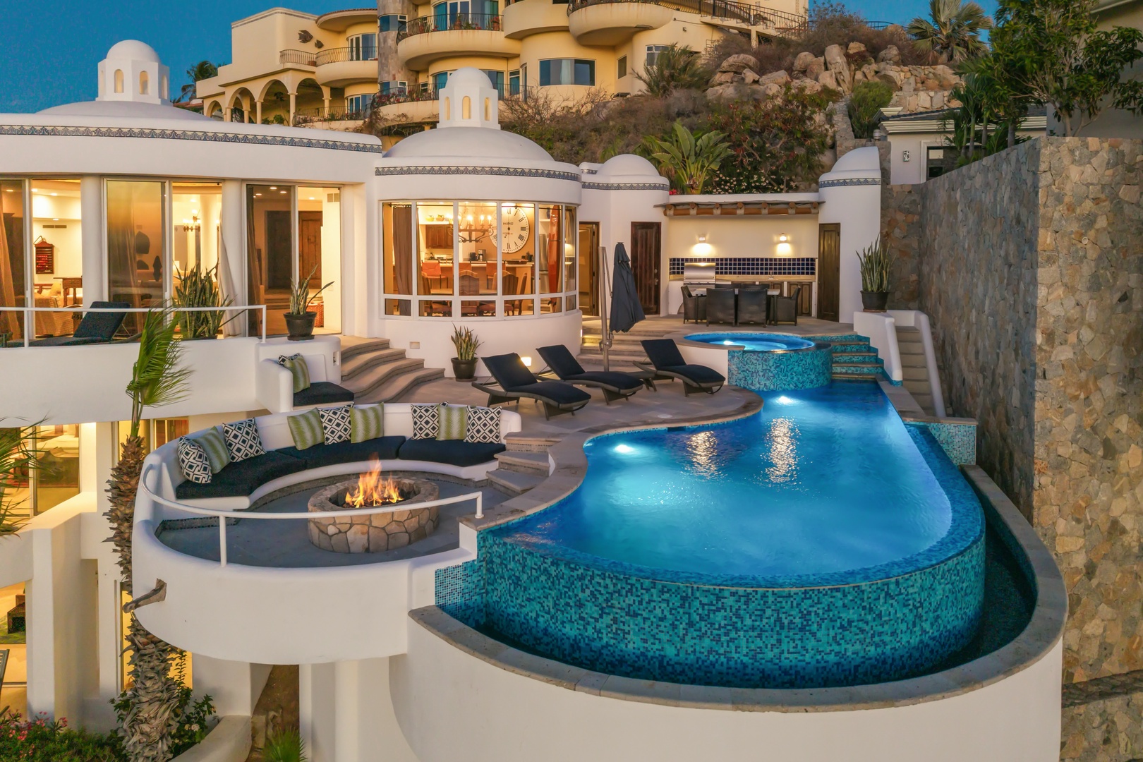 Luxurious hillside villa featuring an infinity pool with integrated spa, outdoor firepit lounge, and stunning architectural design against a dramatic landscape.
