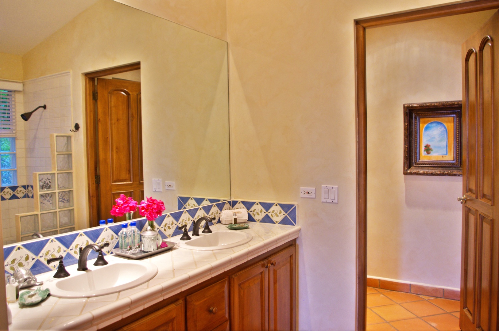 Your en-suite bathroom features dual vanities with handcrafted tile detailing, warm wood cabinetry, and fresh flowers to welcome you. The separate shower area awaits through the glass block privacy wall.