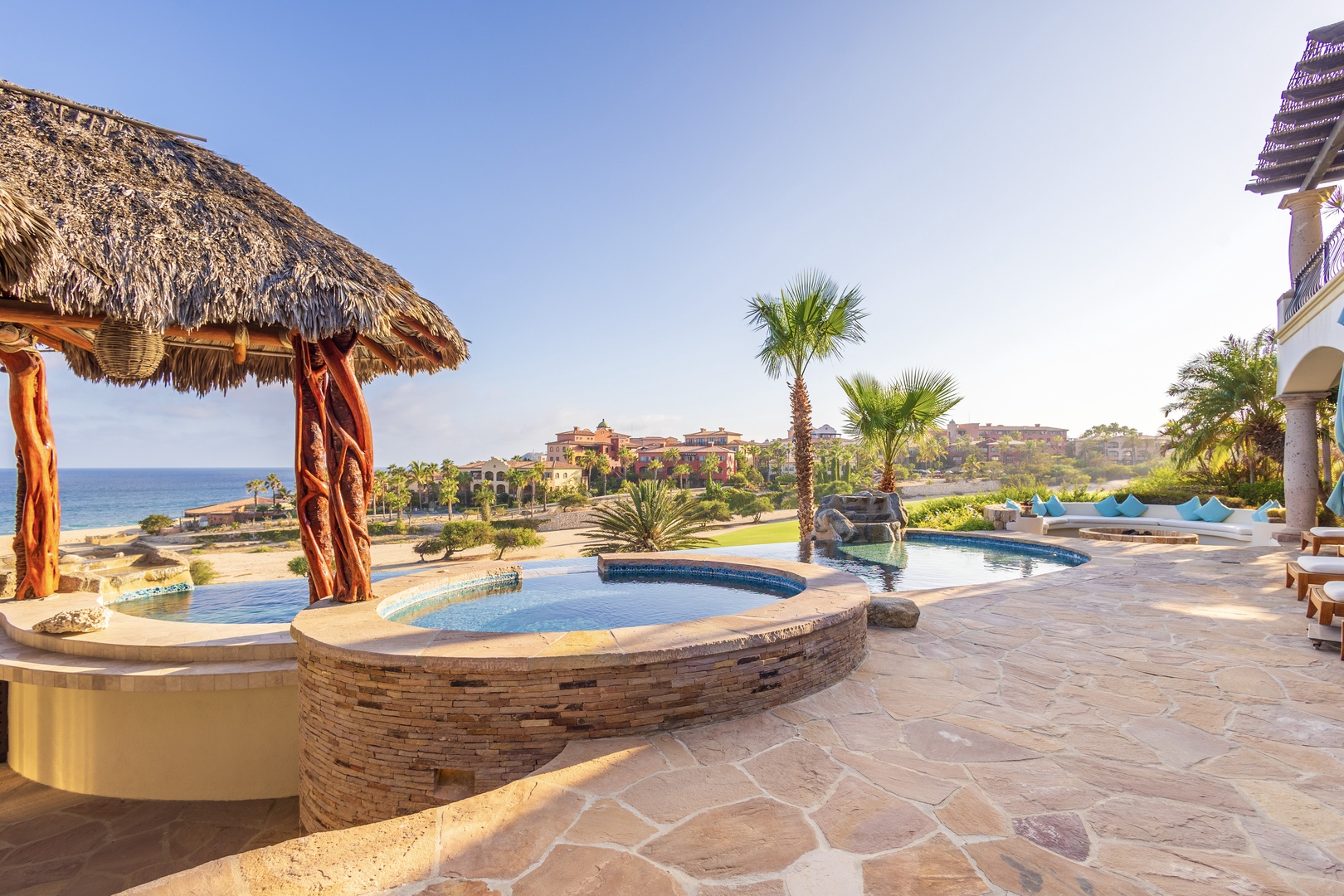 A coastal resort property featuring a multi-level infinity pool area with thatched palapa, palm trees, and beachfront views set against a serene landscape of luxury accommodations.