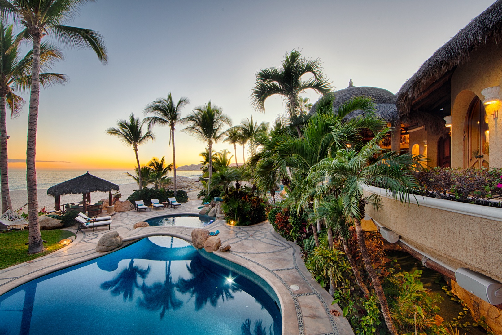 Tropical beachfront villa with private pool at sunset, featuring thatched-roof architecture and lush palm gardens overlooking the ocean.