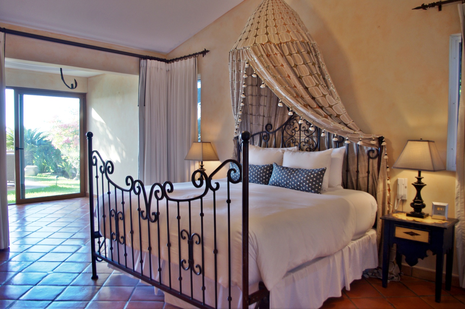 Your romantic bedroom retreat features a stunning canopy draped over an elegant wrought-iron bed, with warm terracotta tiles and lush garden views inviting peaceful relaxation.