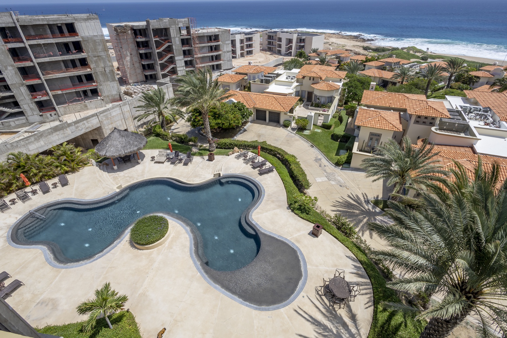 Aerial view of beachfront property showcasing uniquely shaped pool, tropical landscaping, and direct ocean access with nearby residential villas and construction in surrounding neighborhood.