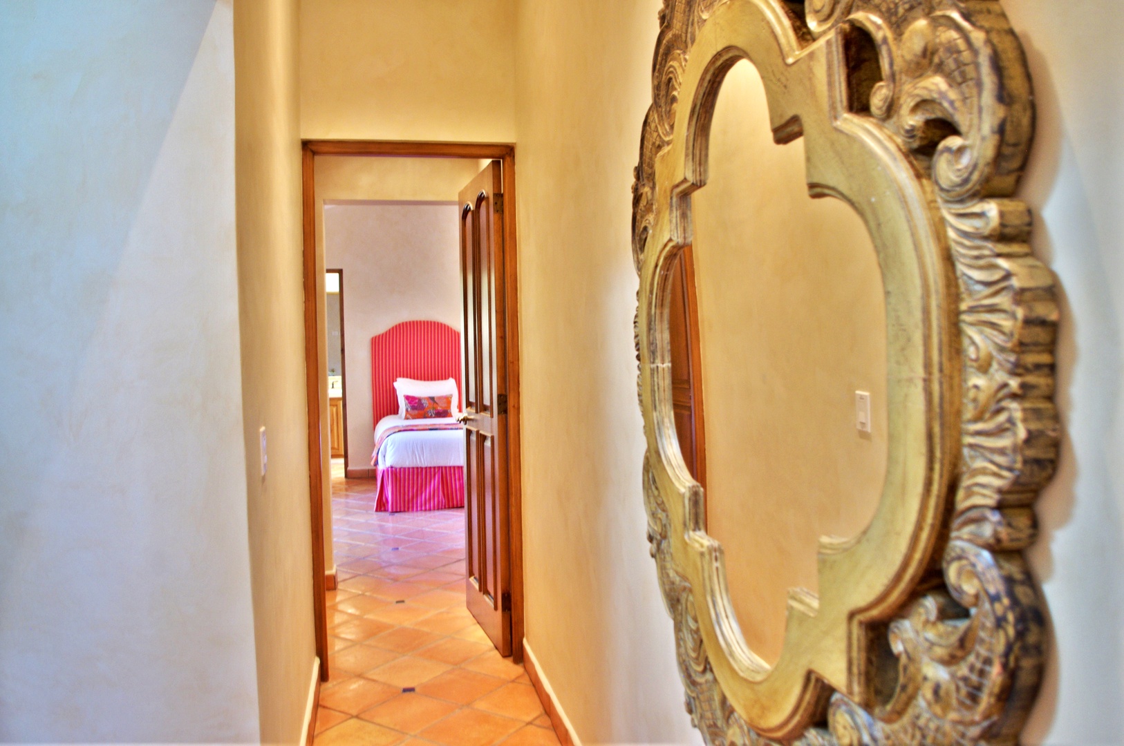 Step through warm terracotta-tiled hallways adorned with ornate golden mirrors leading to your inviting bedroom, where a plush pink bed awaits your arrival.