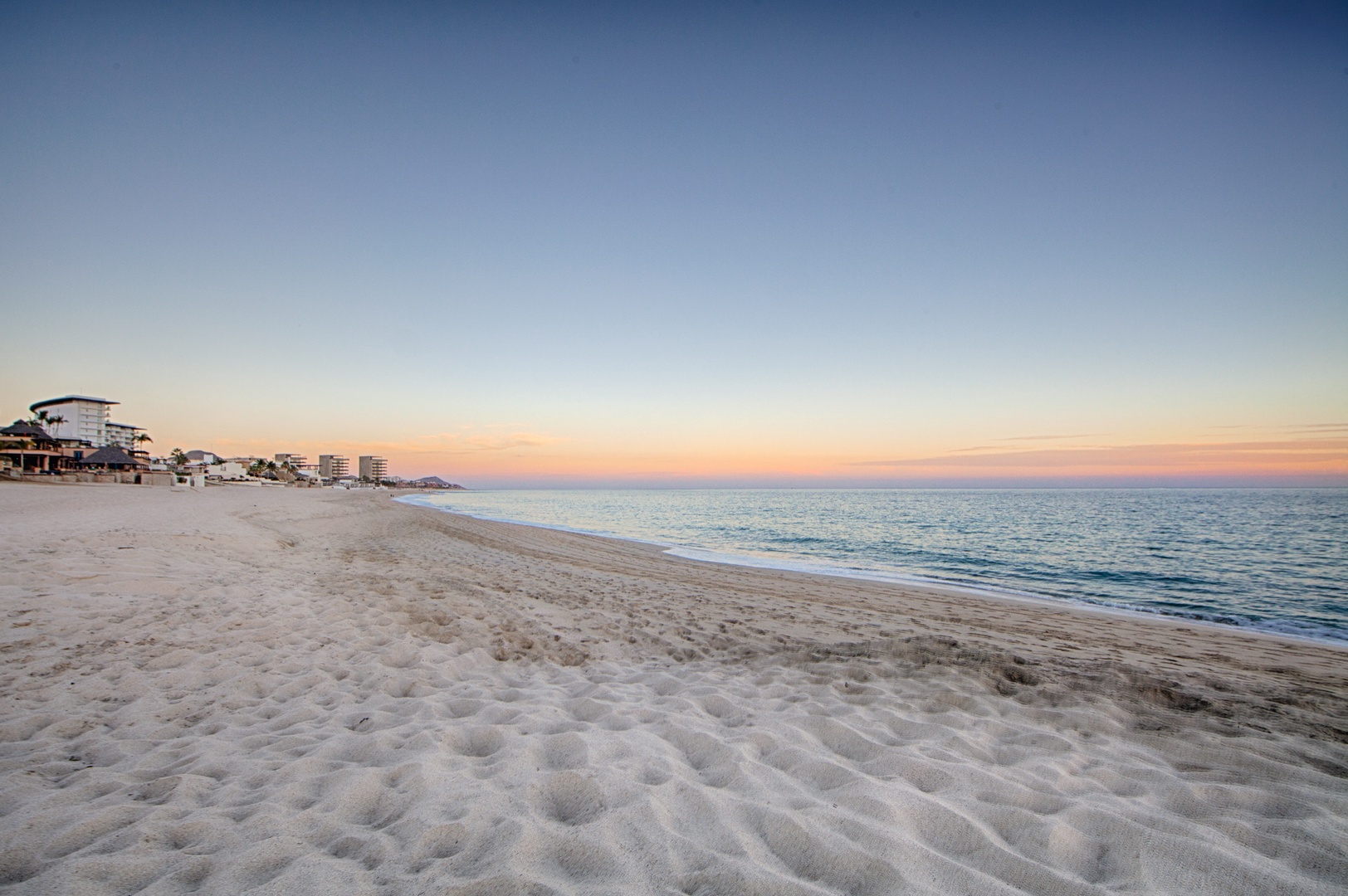 Pristine sandy beach stretches along calm waters at sunset, with coastal buildings dotting the peaceful shoreline.