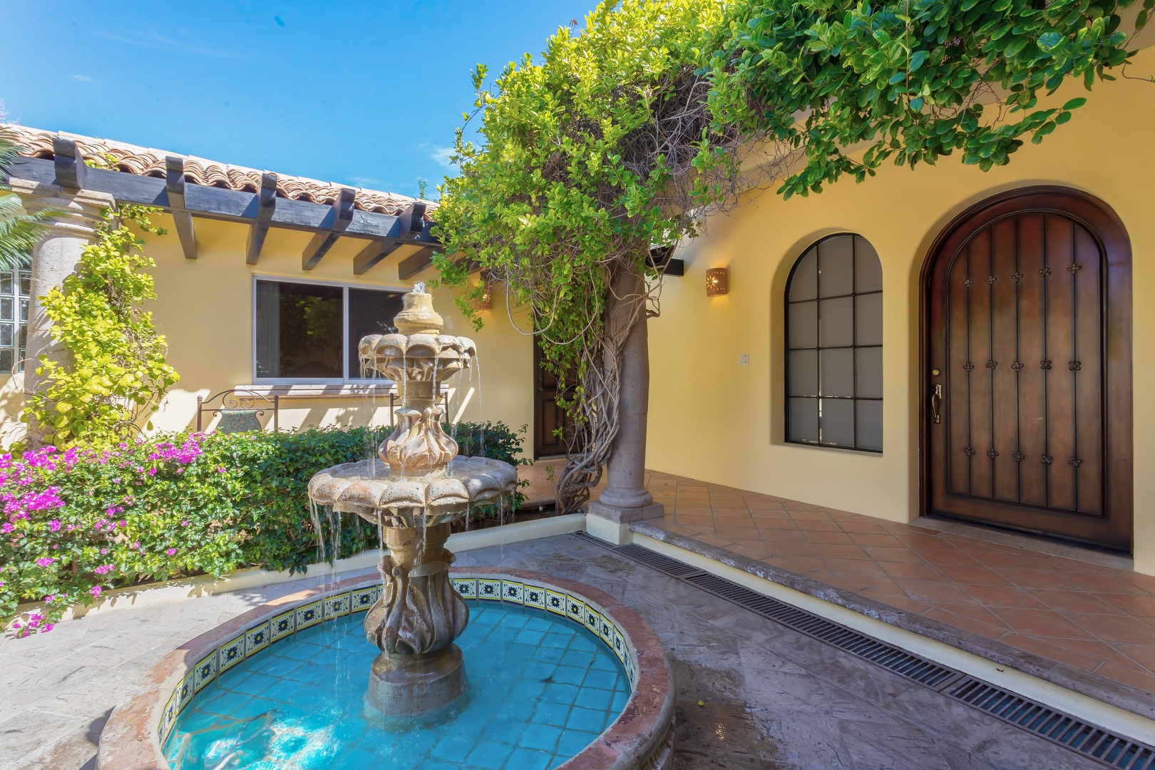 Charming Mediterranean-style property entrance featuring decorative fountain and lush landscaping under clear blue skies.