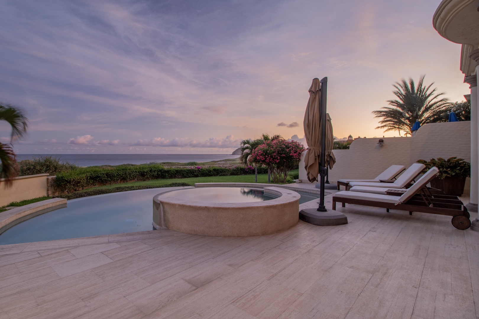 A serene beachfront patio area with pool, jacuzzi, and loungers overlooking the ocean at dusk, surrounded by lush tropical landscaping.
