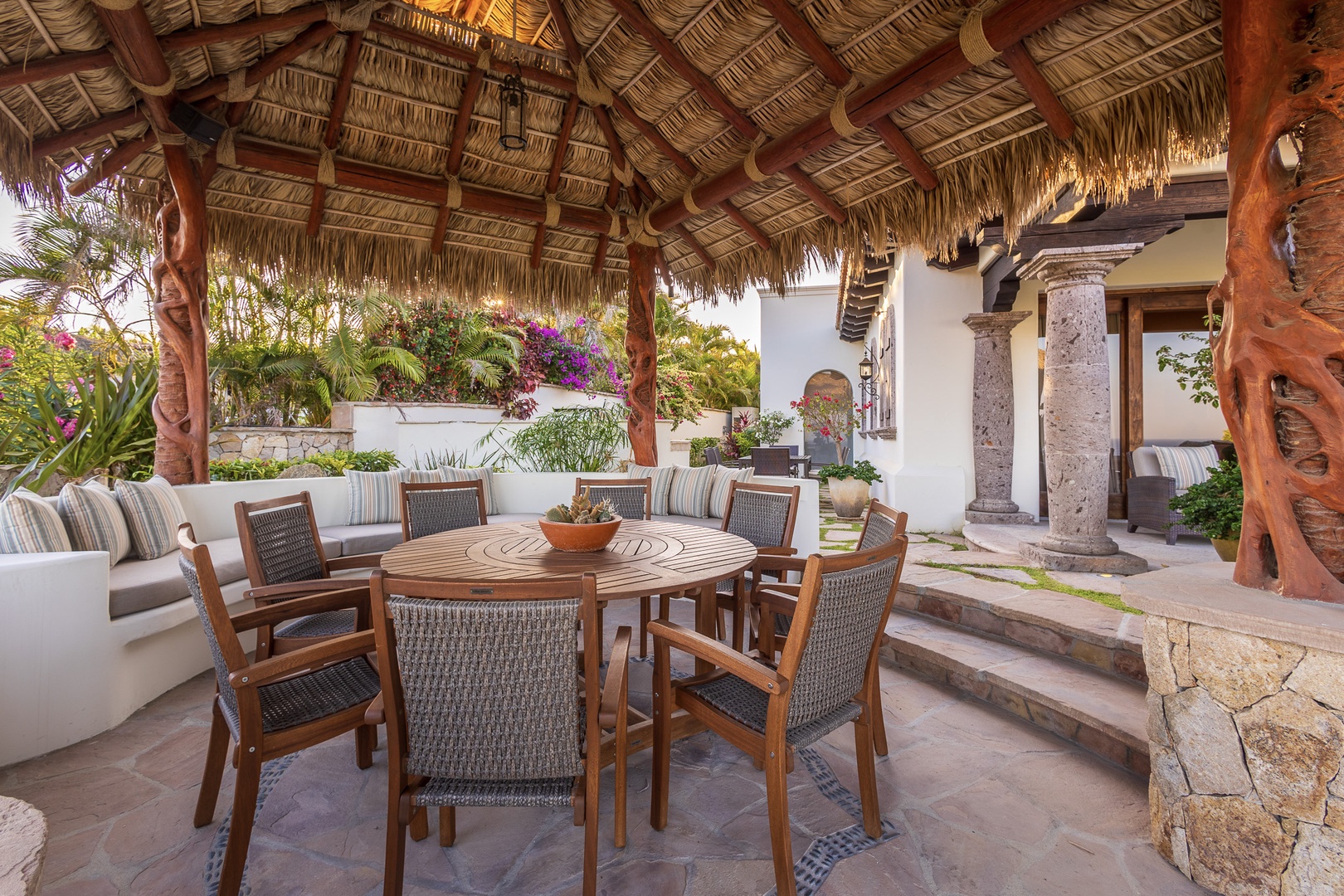 Unwind under the rustic palapa roof at your private outdoor dining area, where a handcrafted wooden table awaits your sun-drenched meals surrounded by tropical greenery and vibrant blooms.