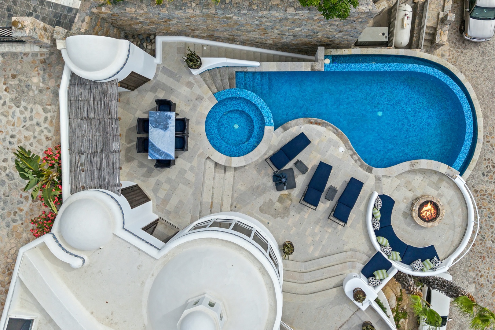Aerial view of a luxurious pool area with curved design, hot tub, outdoor dining spaces, and elegant stone pathways.