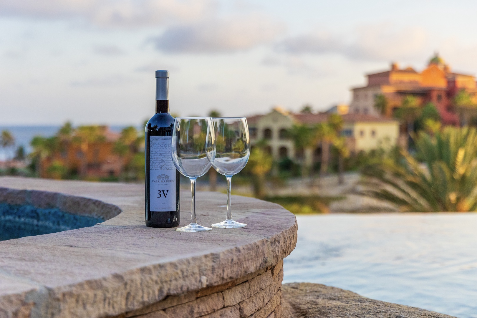 A bottle of wine and two glasses rest on a stone ledge overlooking the resort, with warm architecture and ocean views framing the scene.