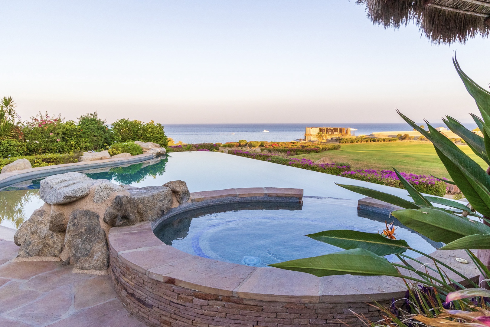 Panoramic coastal views frame an infinity pool and hot tub, surrounded by lush gardens and offering sweeping ocean and fairway vistas.