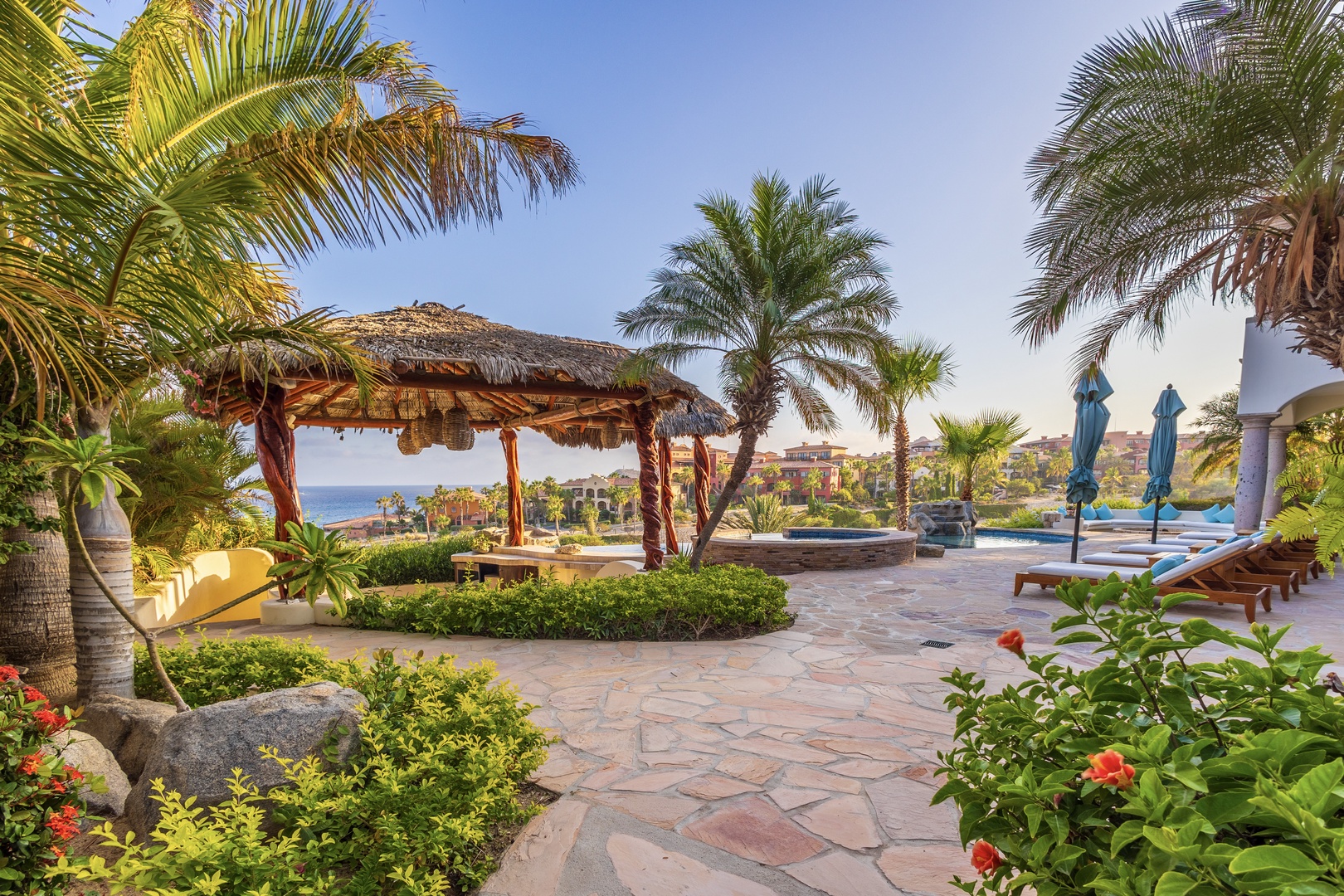 This lush tropical outdoor space features palm-shaded lounging areas, a thatched-roof palapa, and manicured gardens overlooking the ocean and nearby resort.