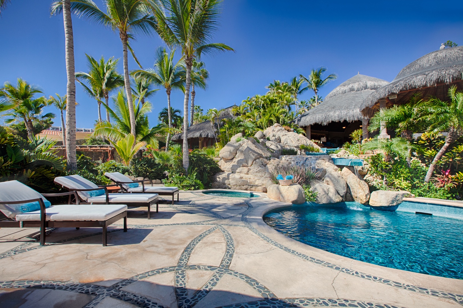 Tropical paradise pool complex with natural rock waterfall features and traditional thatched-roof buildings.